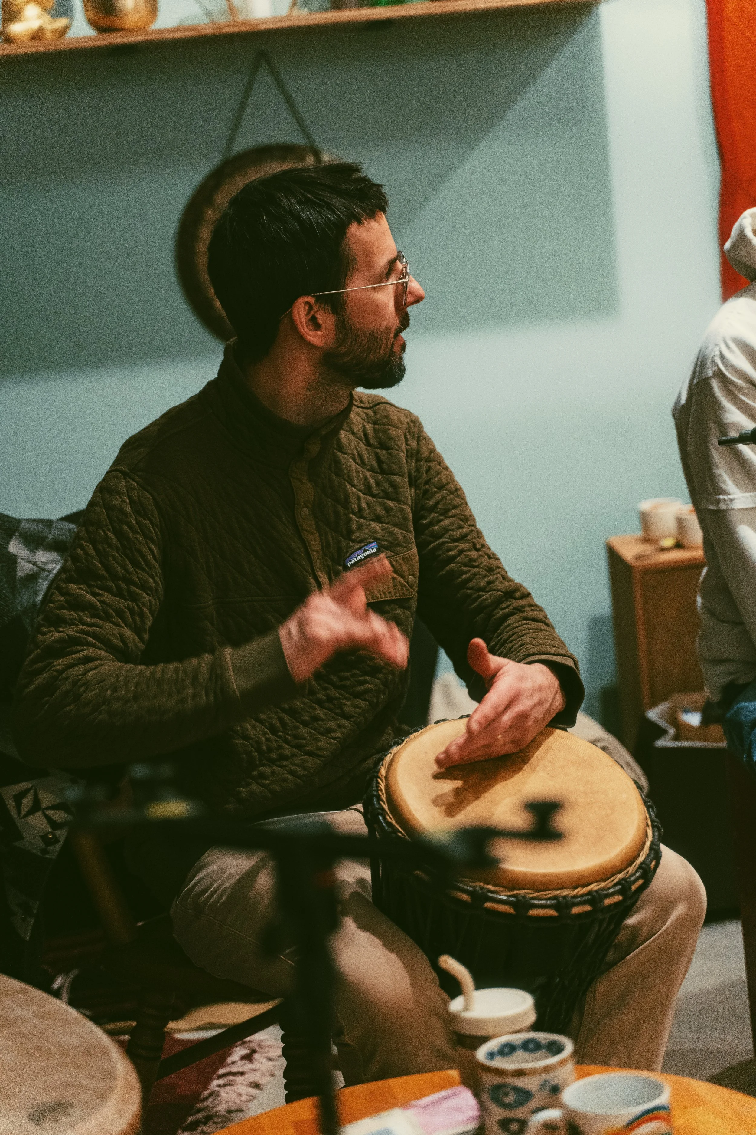 A man with glasses and a beard playing a drum indoors, seated at a table with cups and other items nearby.