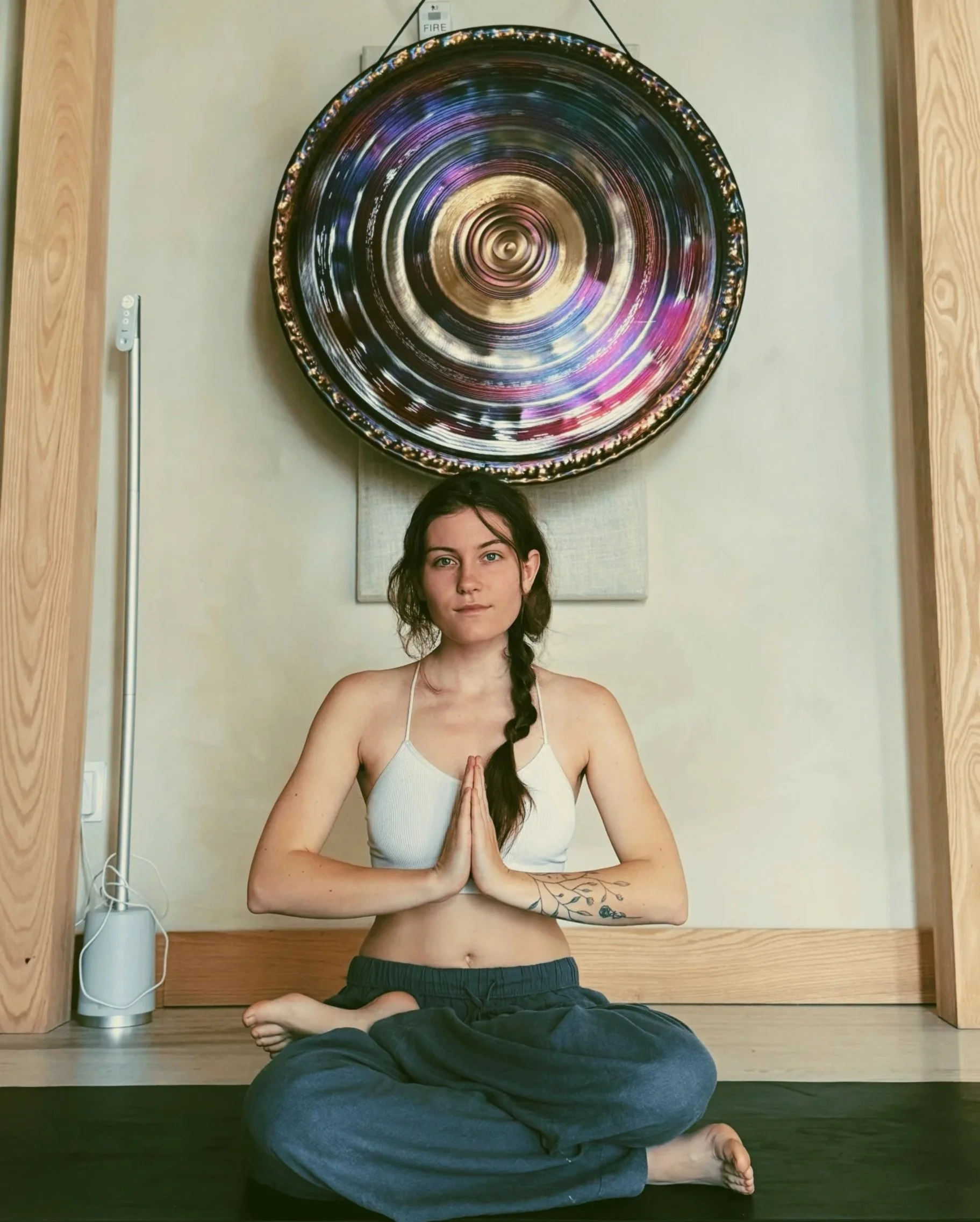 A young woman practicing meditation indoors, sitting cross-legged on a black mat with her hands in prayer position, in front of a circular abstract art piece hanging on the wall.