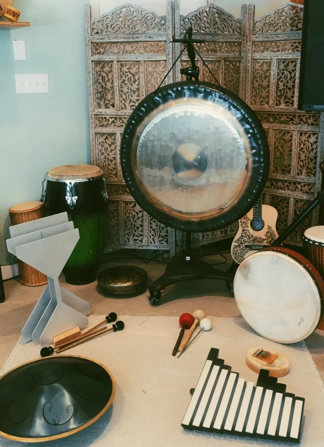 A collection of musical instruments including a large gong, djembe, bongos, chimes, a tambourine, and a hammer and mallets for percussion, arranged in a room with decorative wooden room dividers in the background.