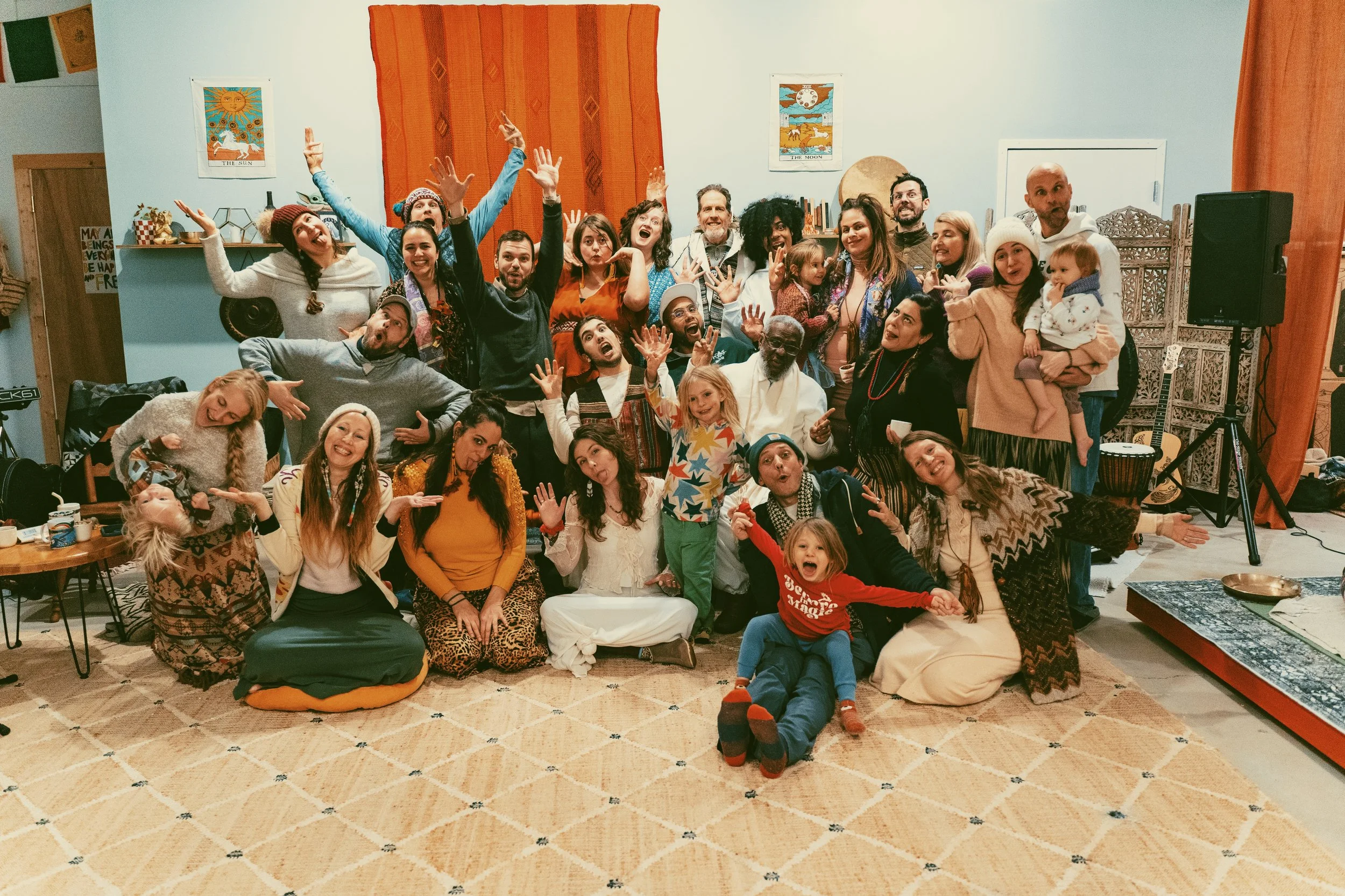 A large group of people, including children and adults, posing together indoors in a cozy, decorated room, smiling and making playful gestures.
