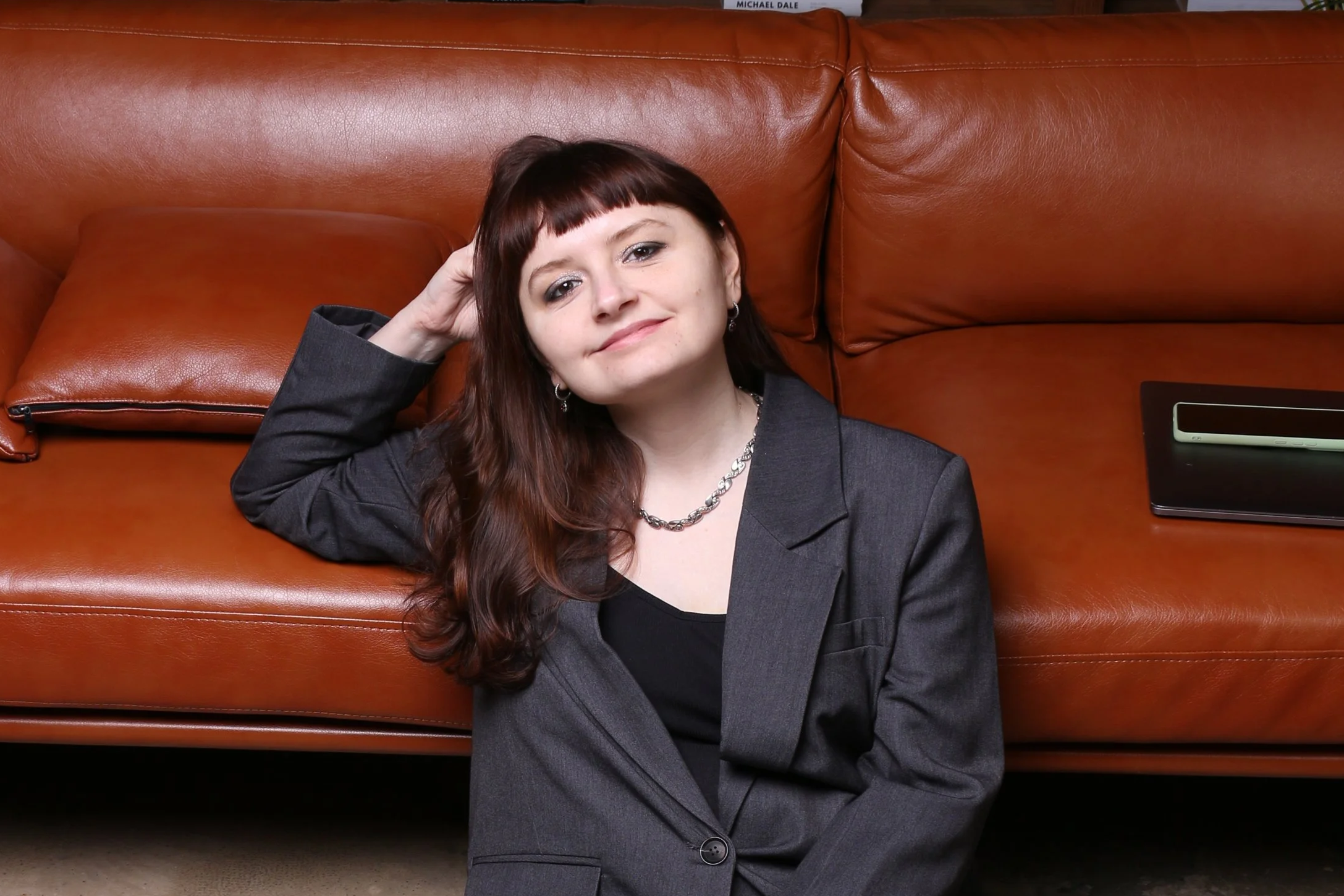 A woman with shoulder-length brown hair, bangs, and light skin is sitting on the floor, leaning against a large brown leather sofa. She is wearing a gray blazer, a black top, earrings, and a necklace. She is smiling lightly and has her right hand behind her head. A piece of black and beige electronic device is on the sofa next to a closed black laptop.