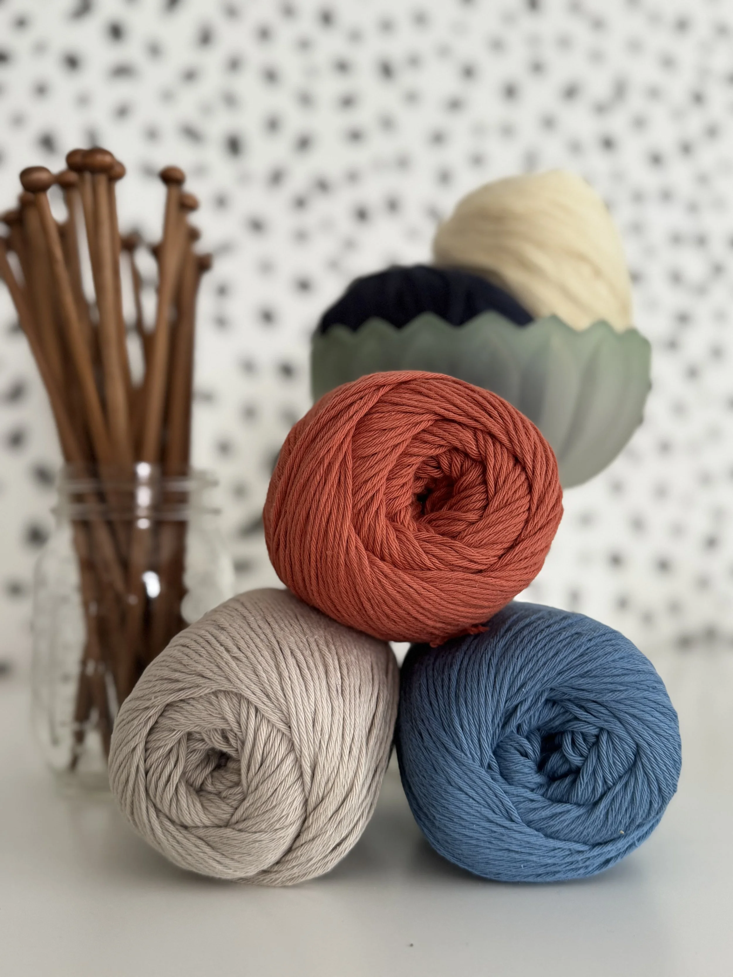 Three balls of yarn in red, beige, and blue, stacked on a white surface, with a jar of knitting needles behind them and a decorative bowl with yarn in the background.