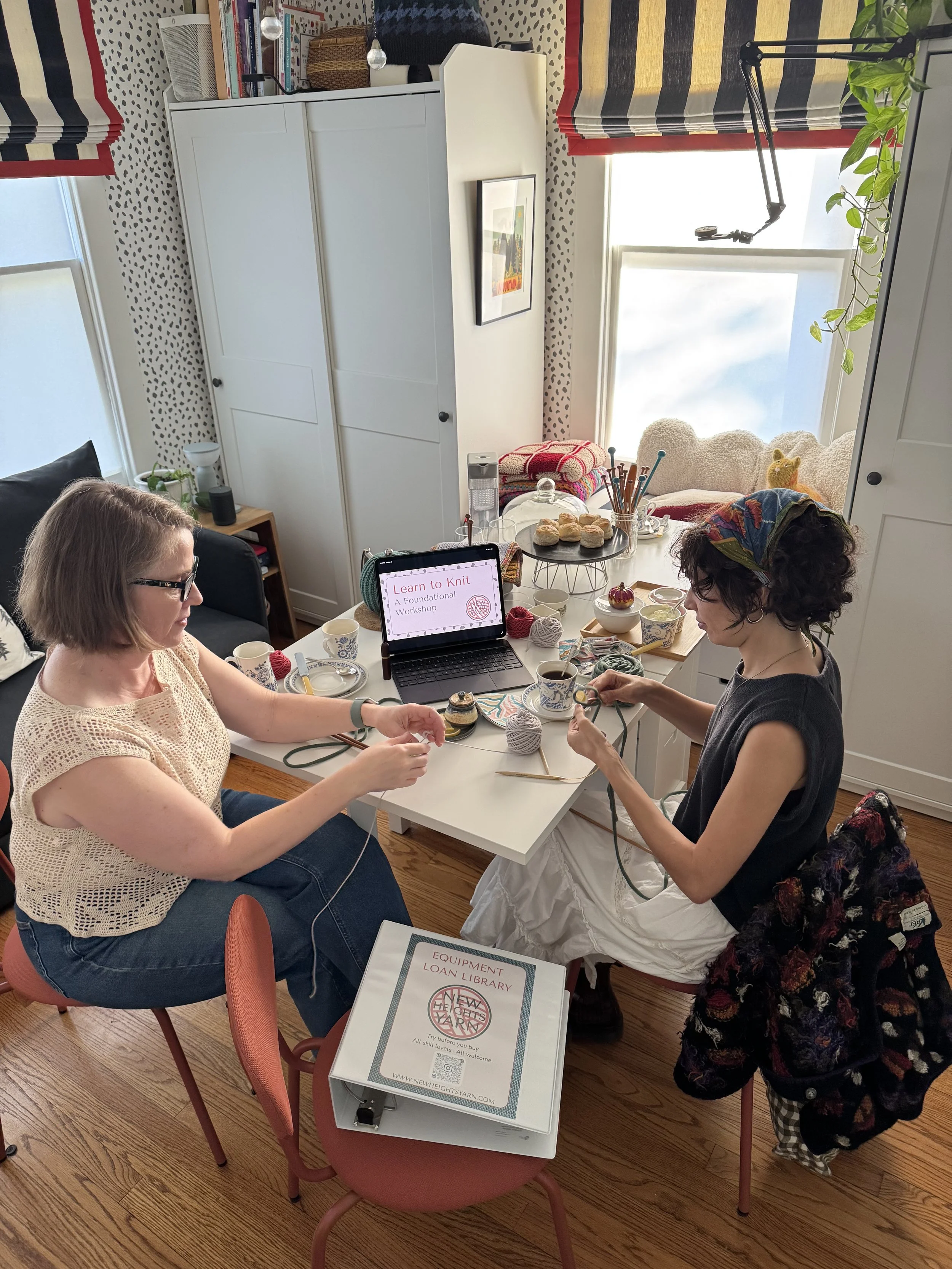 Two women knitting at a table in a cozy, well-lit room with a laptop displaying 'Learn to Knit: A Foundational Workshop,' surrounded by craft supplies, cups, and baked goods.