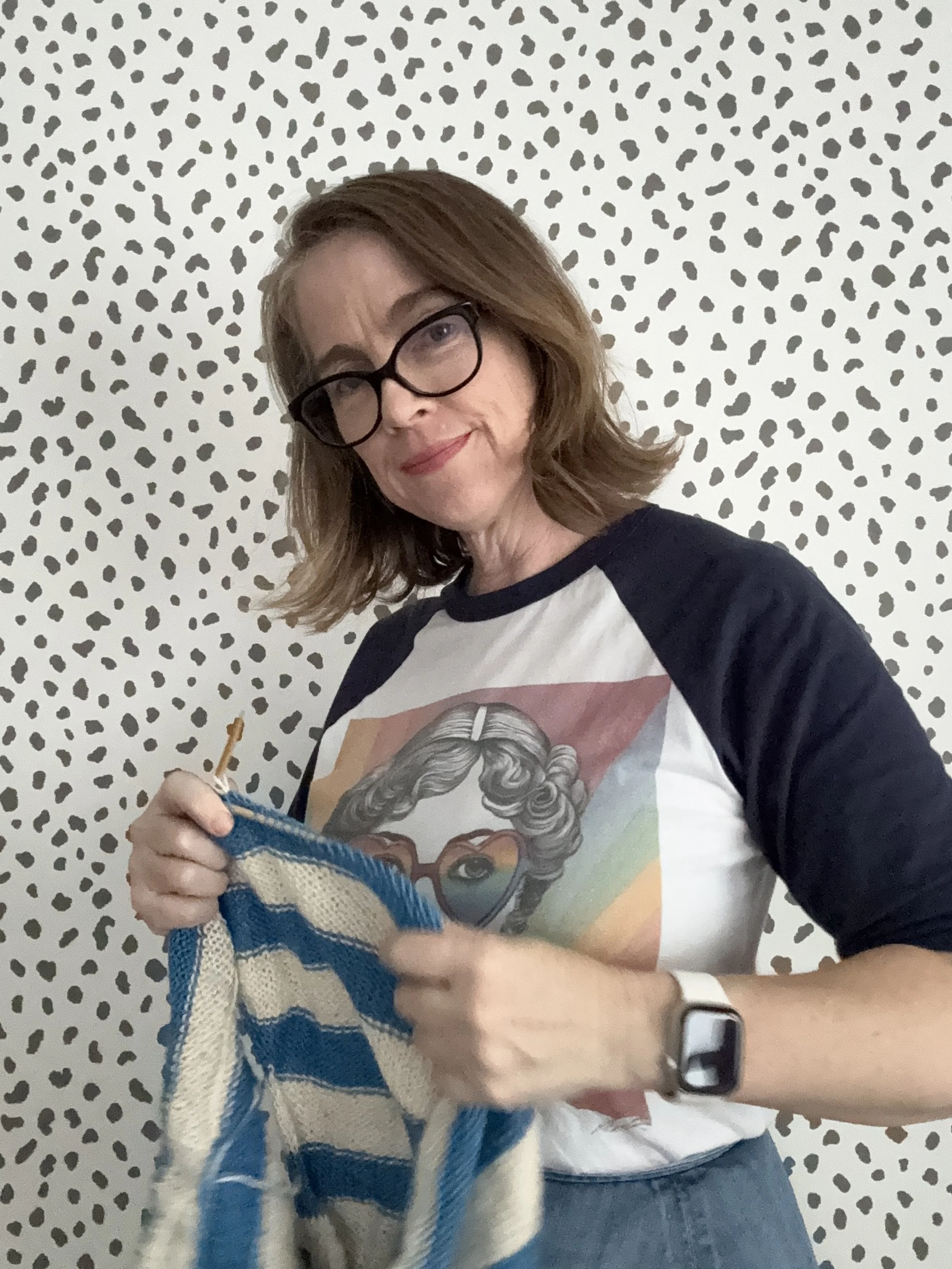 Woman with glasses knitting a striped blue and white garment in front of a black and white dotted patterned wall.