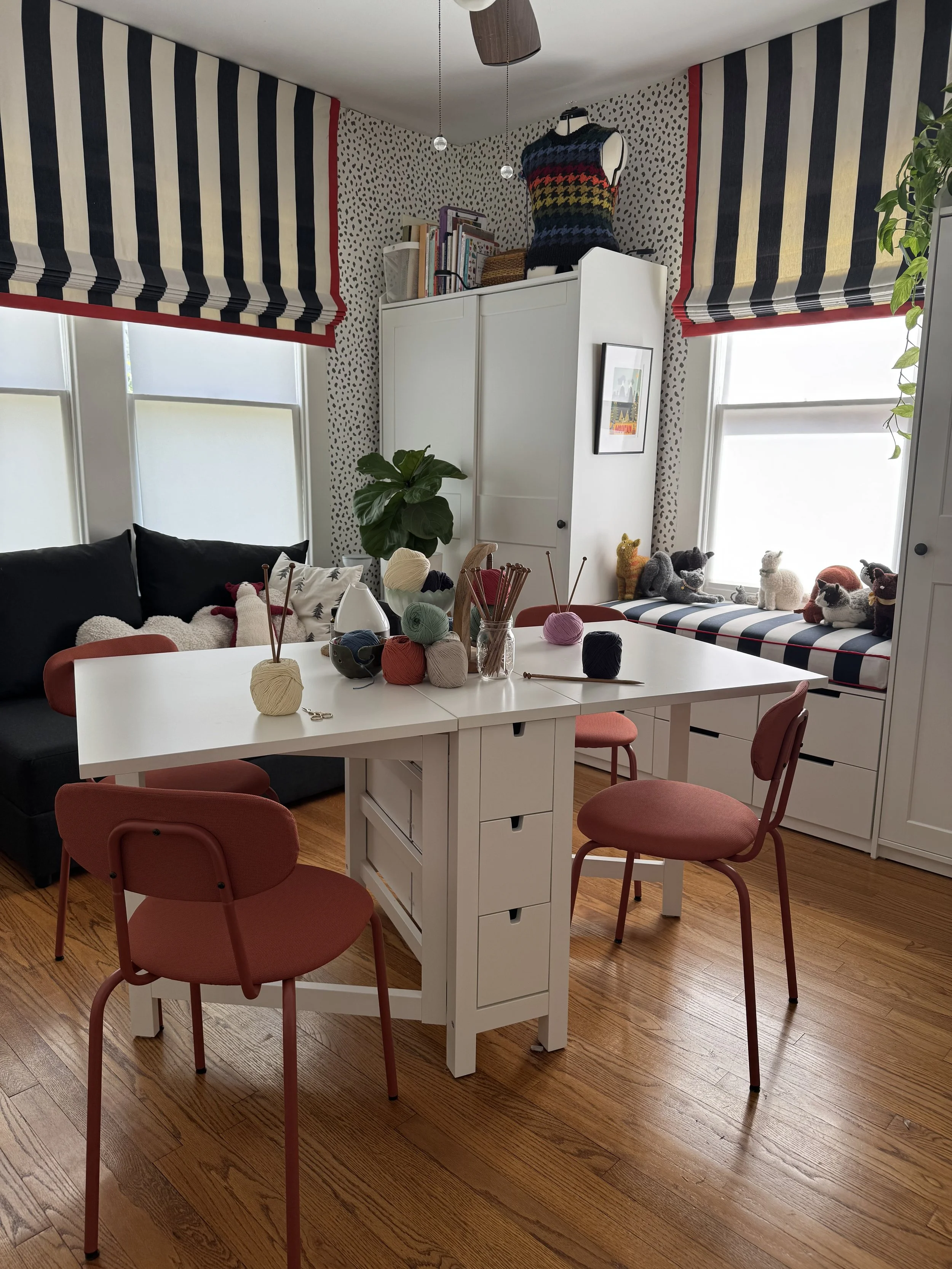 A cozy dining area with a white table, pink chairs, and a window seat with plush toy animals. The room has black and white patterned wallpaper, striped window treatments, and knit yarn balls on the table.