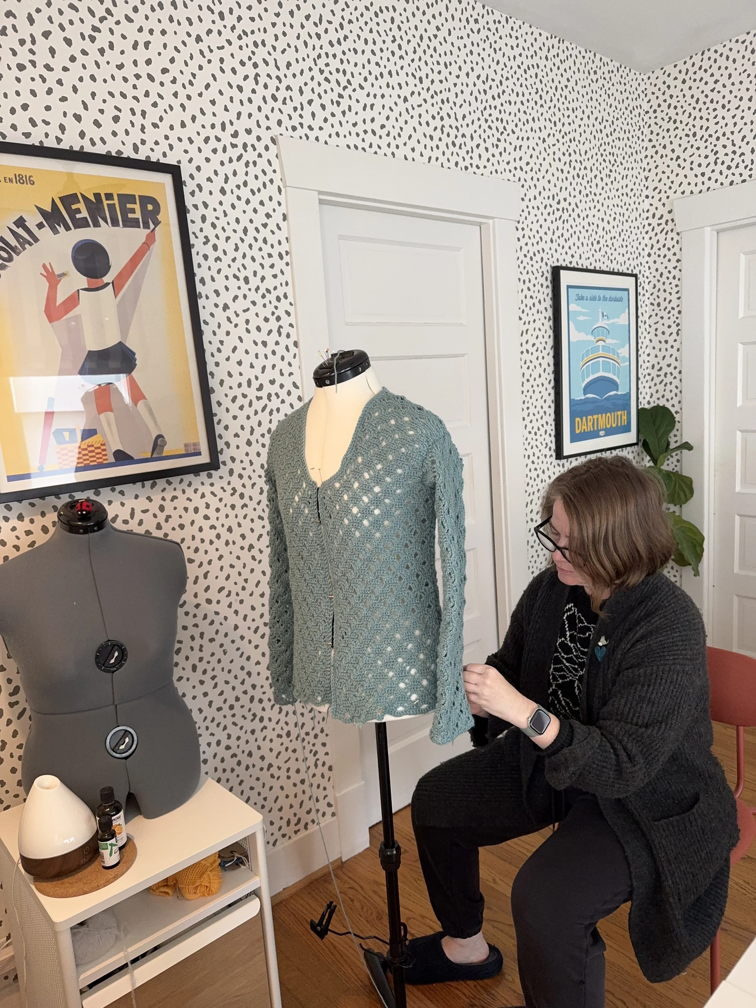Woman working on a mannequin dressed in a crochet sweater in a room with spotted wallpaper and framed art on the walls.
