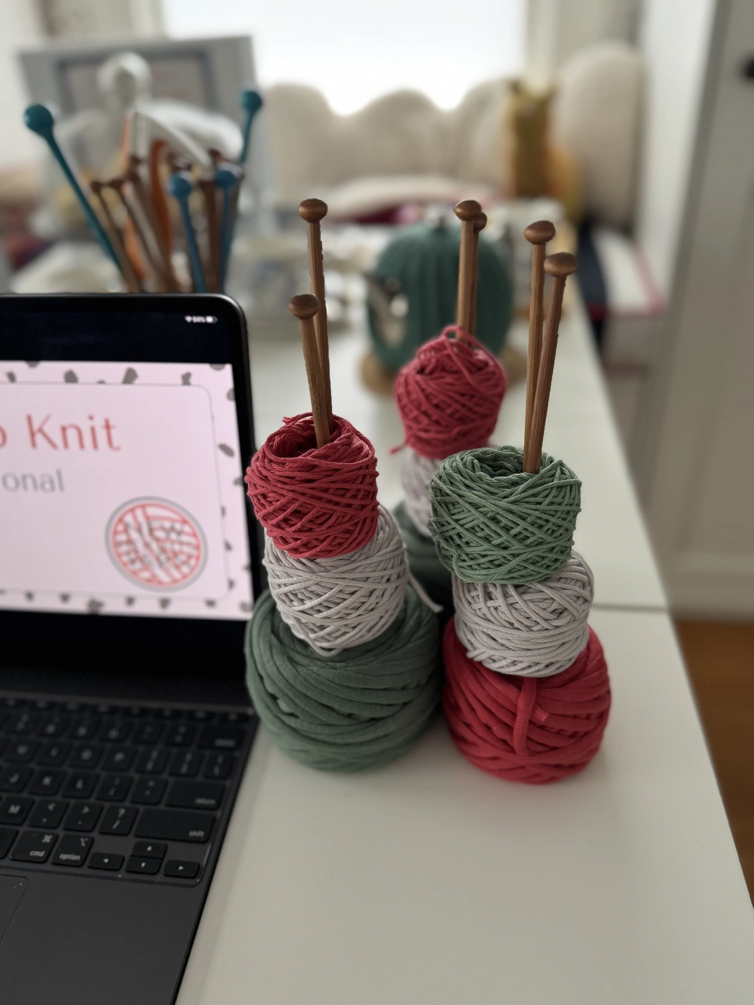 Multiple balls of yarn in red, green, and beige colors stacked with knitting needles inserted into some of them on a white surface. A laptop and a sign with a knitting logo are also visible.
