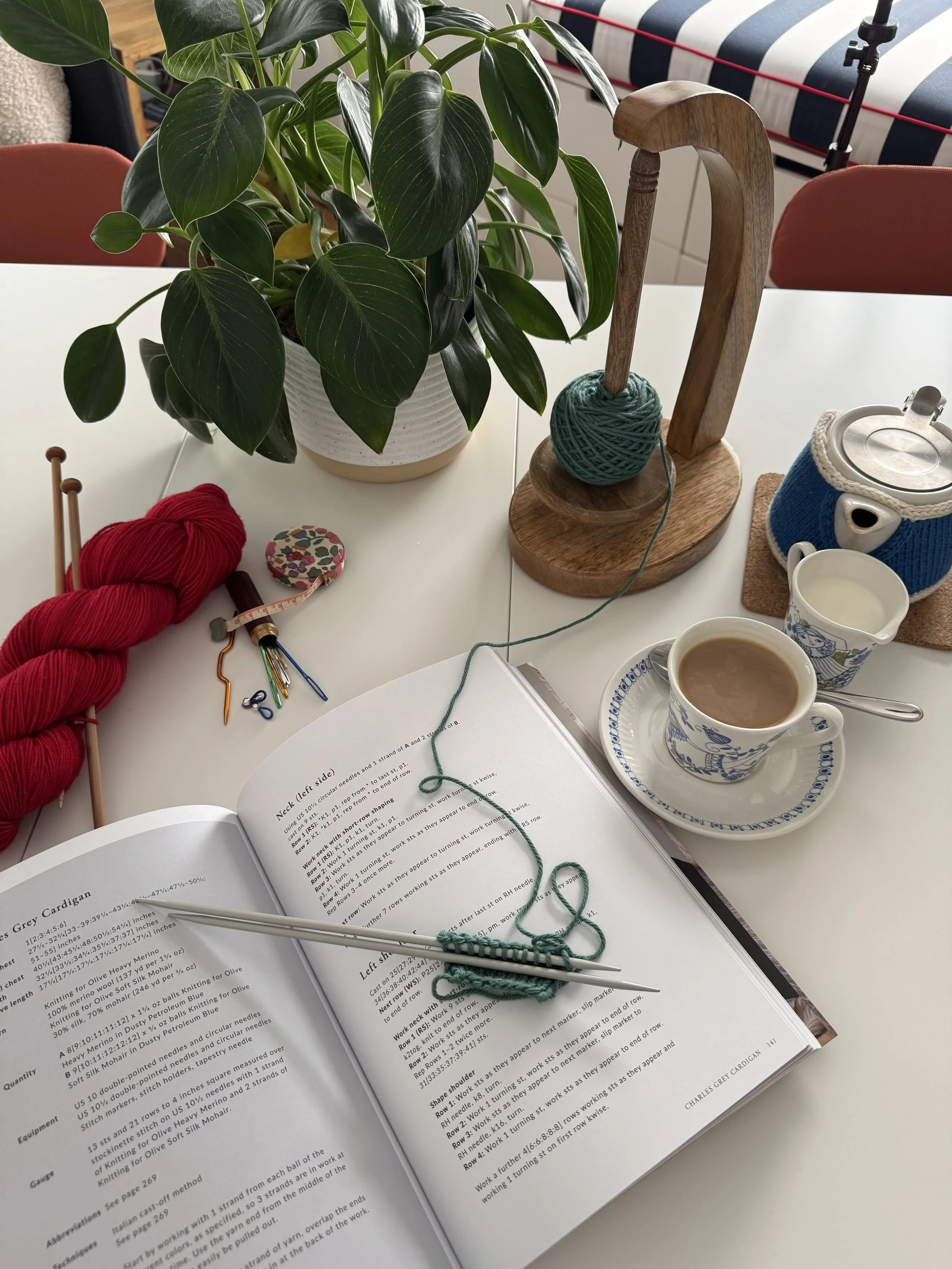 A table with a potted plant, a ball of yarn with knitting needles, an open knitting book, a cup of coffee, and a teapot.