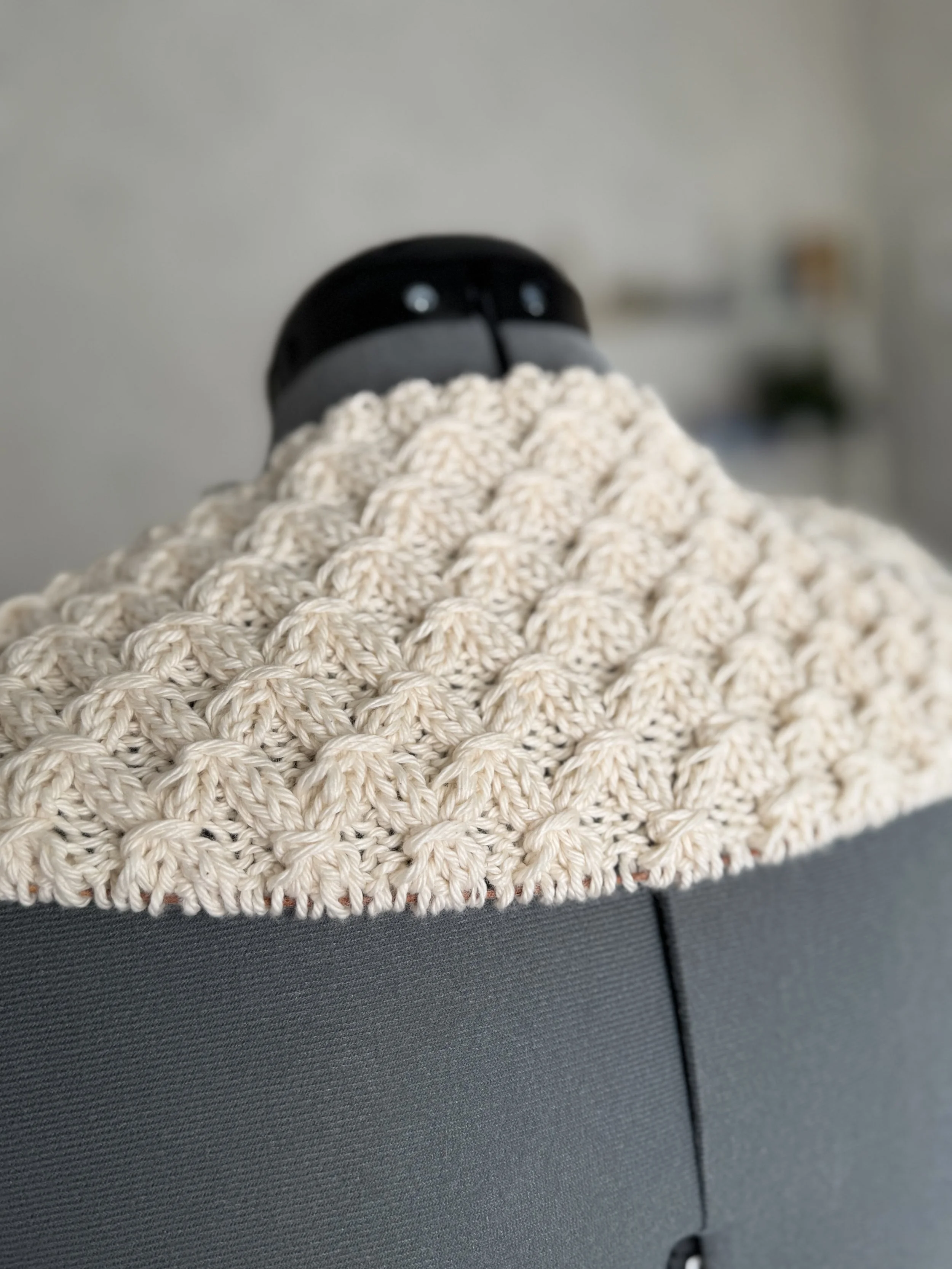 A black robot vacuum cleaner covered with a beige, chunky knit blanket sitting on a grey couch.