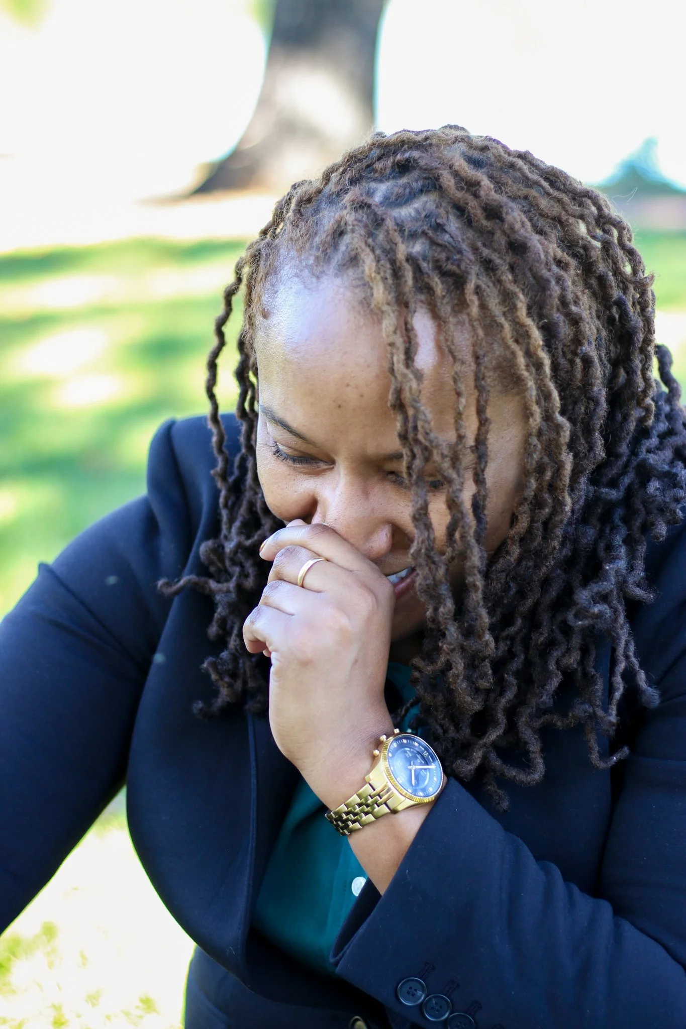 A woman with long curly hair in a ponytail, wearing a Yamaha watch, a dark blazer, and a green shirt, smiling outdoors with her hand near her face.