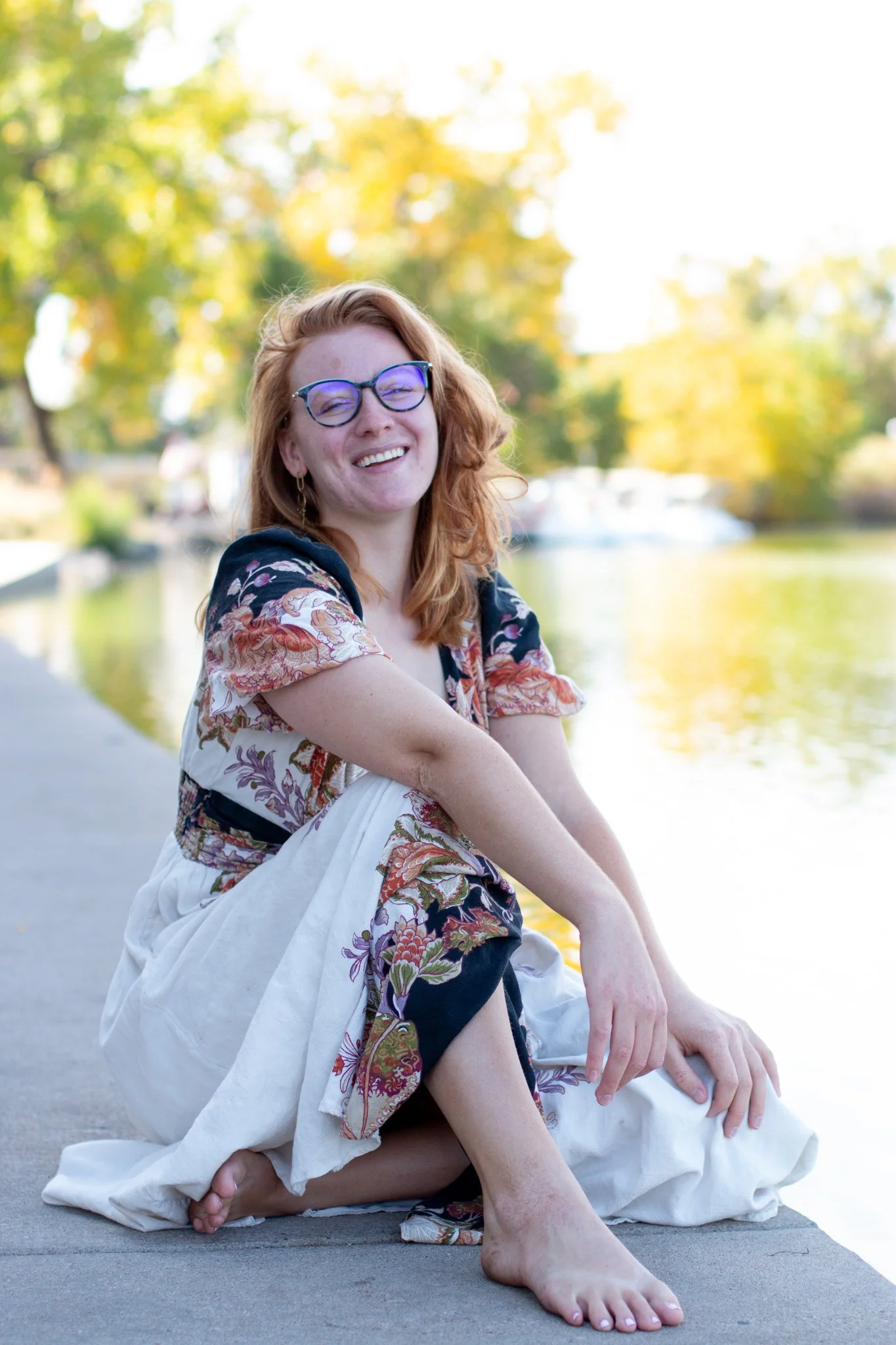 A young woman with red hair and glasses sitting barefoot on a concrete path by a lake, smiling at the camera with trees and water in the background.