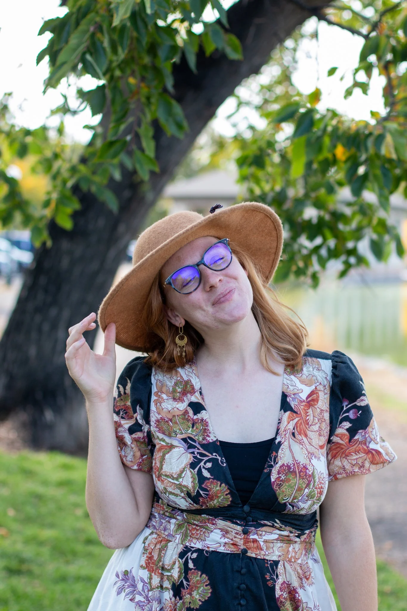 A woman with red hair, wearing glasses, earrings, a tan wide-brimmed hat, and a floral dress, standing outdoors near a tree, with her hand touching her hat and a slight smile.