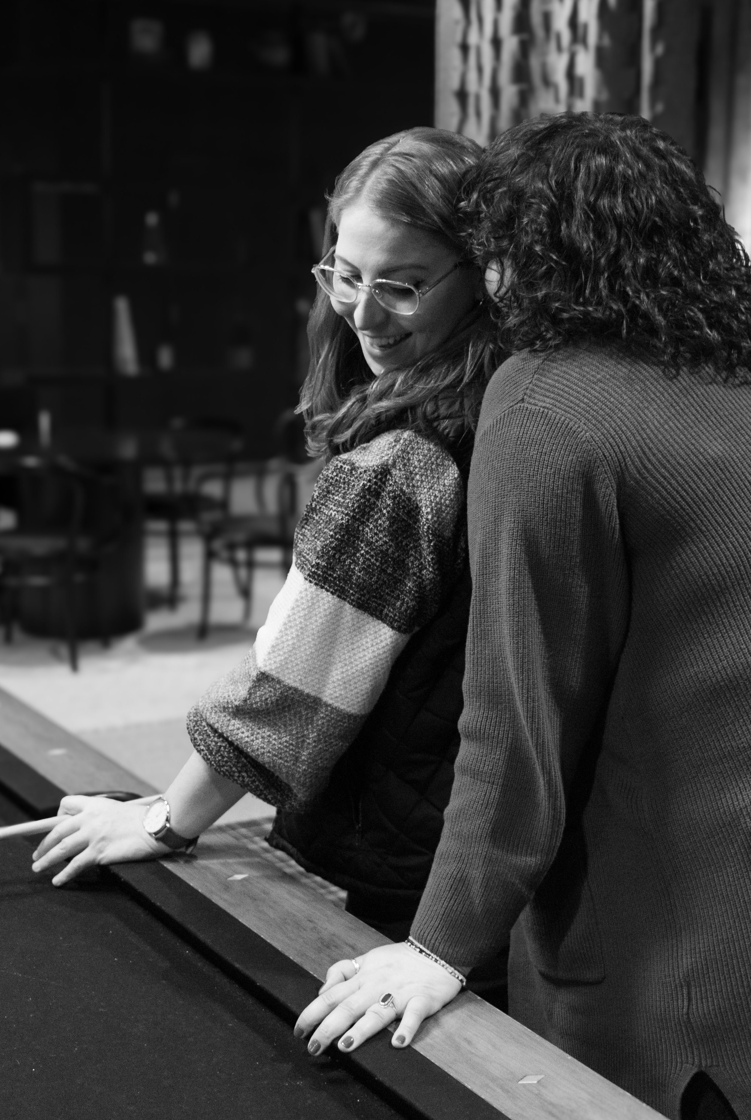 Two women leaning over a pool table, smiling and engaging in conversation, in a room with chairs and shelving in the background.