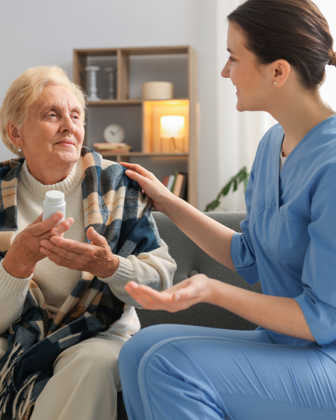 A healthcare worker in blue scrubs talking to an elderly woman in a cozy living room.