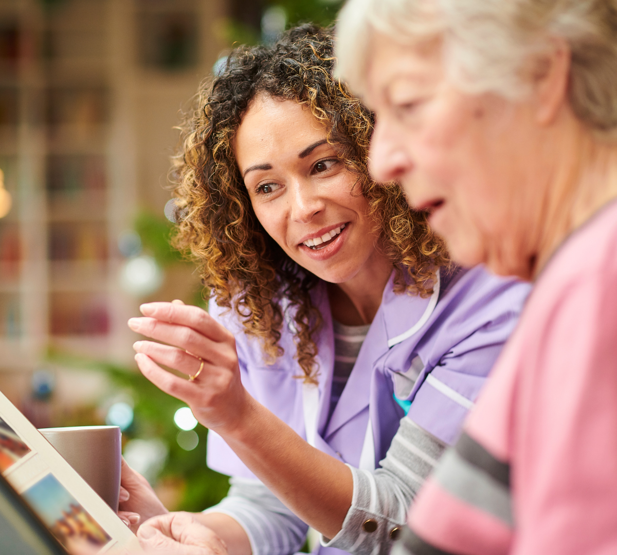 A young female healthcare worker with curly hair, wearing a purple medical uniform, is talking to an elderly woman with short gray hair, who is wearing a pink shirt, in a bright, cozy setting.