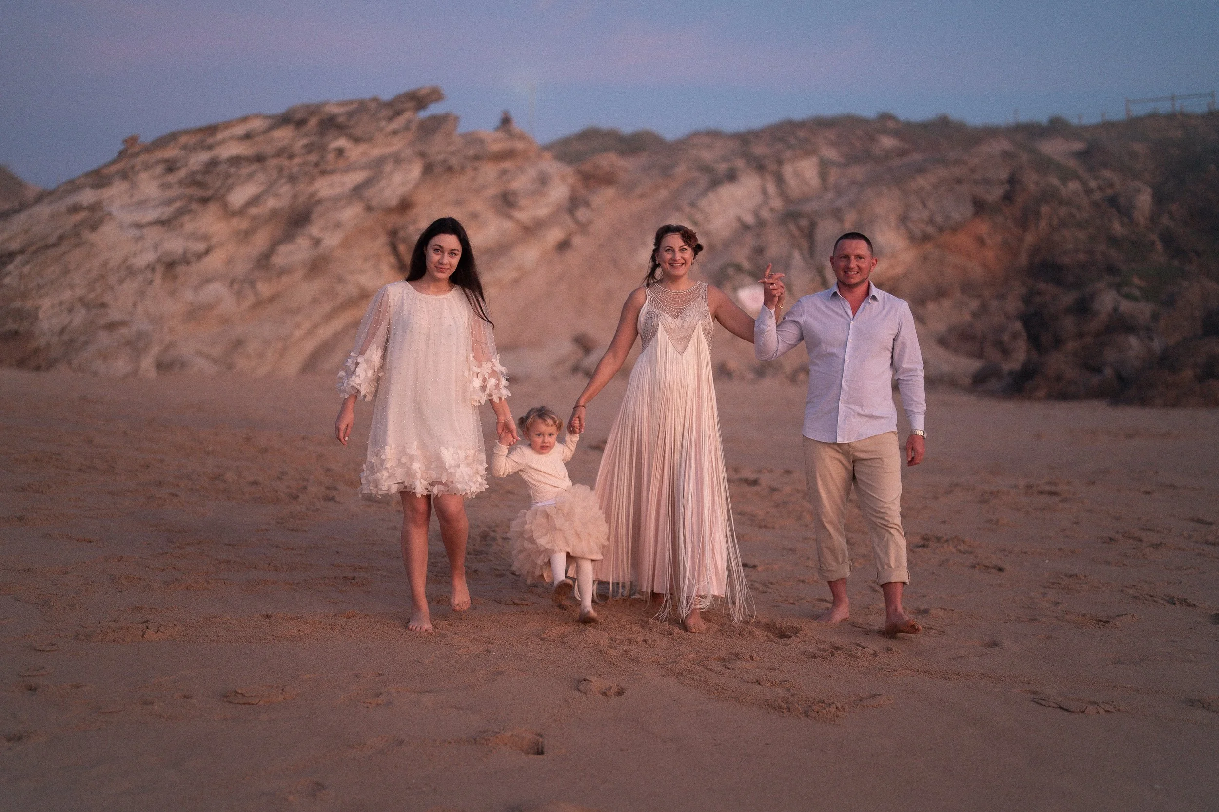 A family of four walking on a beach during sunset, with rocky cliffs in the background. The mother and daughter are holding hands, the daughter is in a cream-colored dress, and the father is holding the mother's hand. The family appears joyful, dressed in light-colored, semi-formal clothing, with the scene illuminated by soft, warm light.