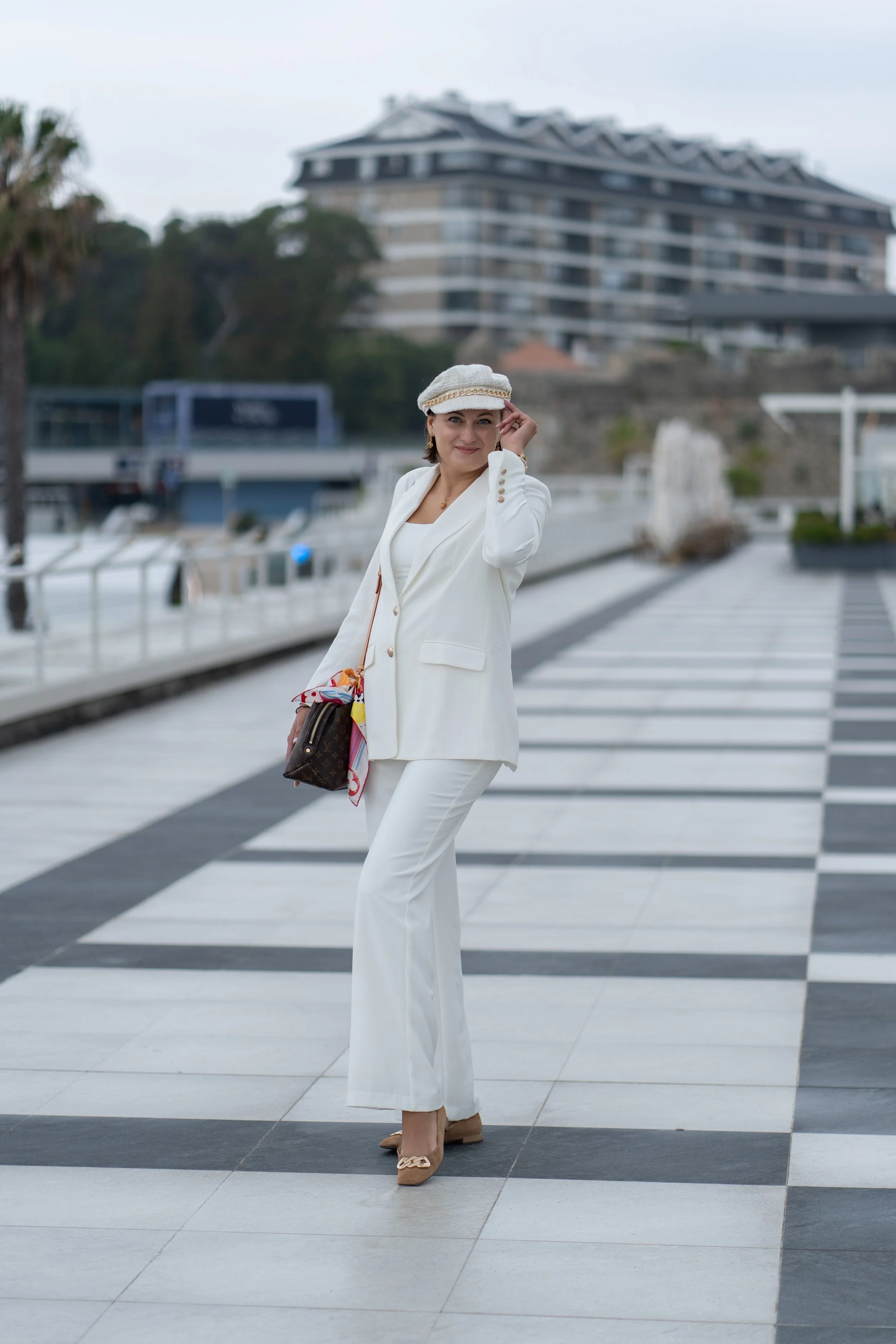 A woman dressed in an all-white suit, wearing a white hat and beige shoes, standing on a checkered promenade near water with city buildings in the background.