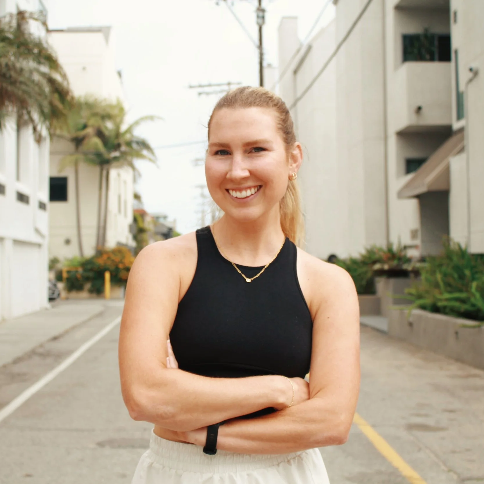 Smiling woman with blonde hair in a ponytail, wearing a black sleeveless top and light-colored pants, standing outdoors with arms crossed on a city street.