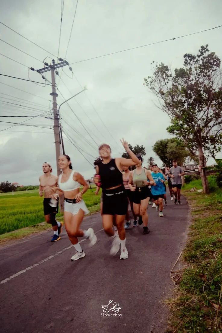 Group of people running on a rural road, some are shirtless, with trees and power lines overhead, and cloudy sky