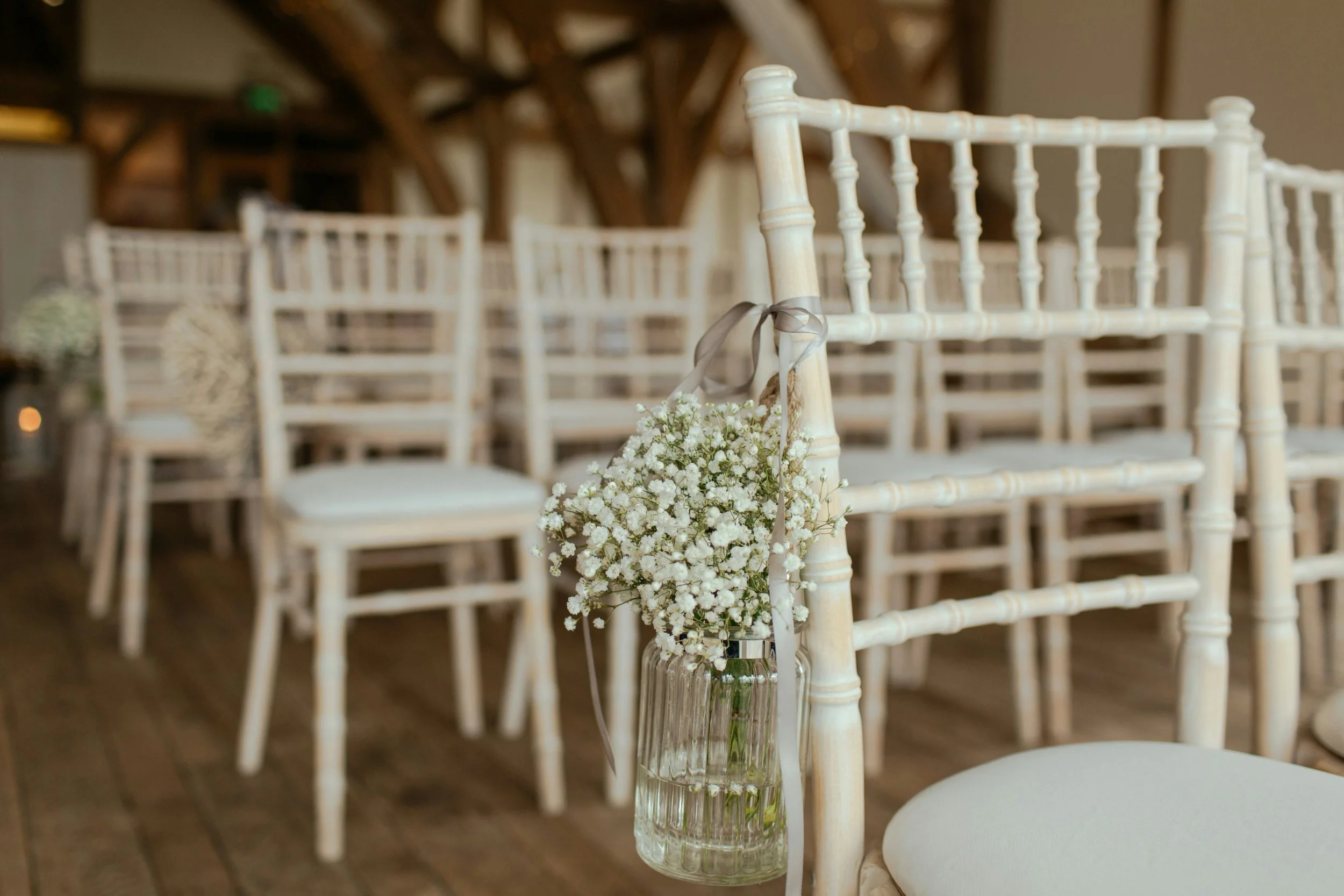 White chairs with a flower bouquet hanging on the corner of one chair in an indoor venue, likely set up for a wedding or event.