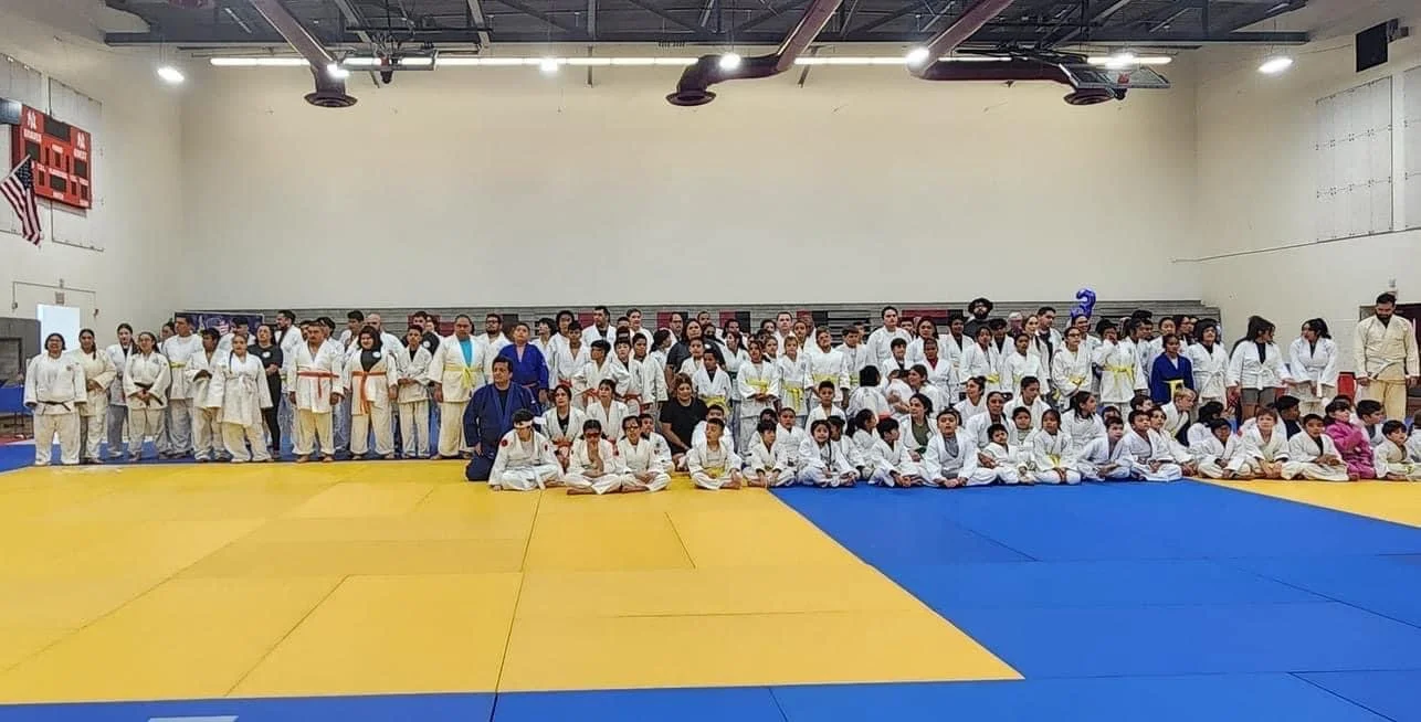 Group of children and adults in judo uniforms sitting and standing on a yellow and blue mat in a gymnasium.