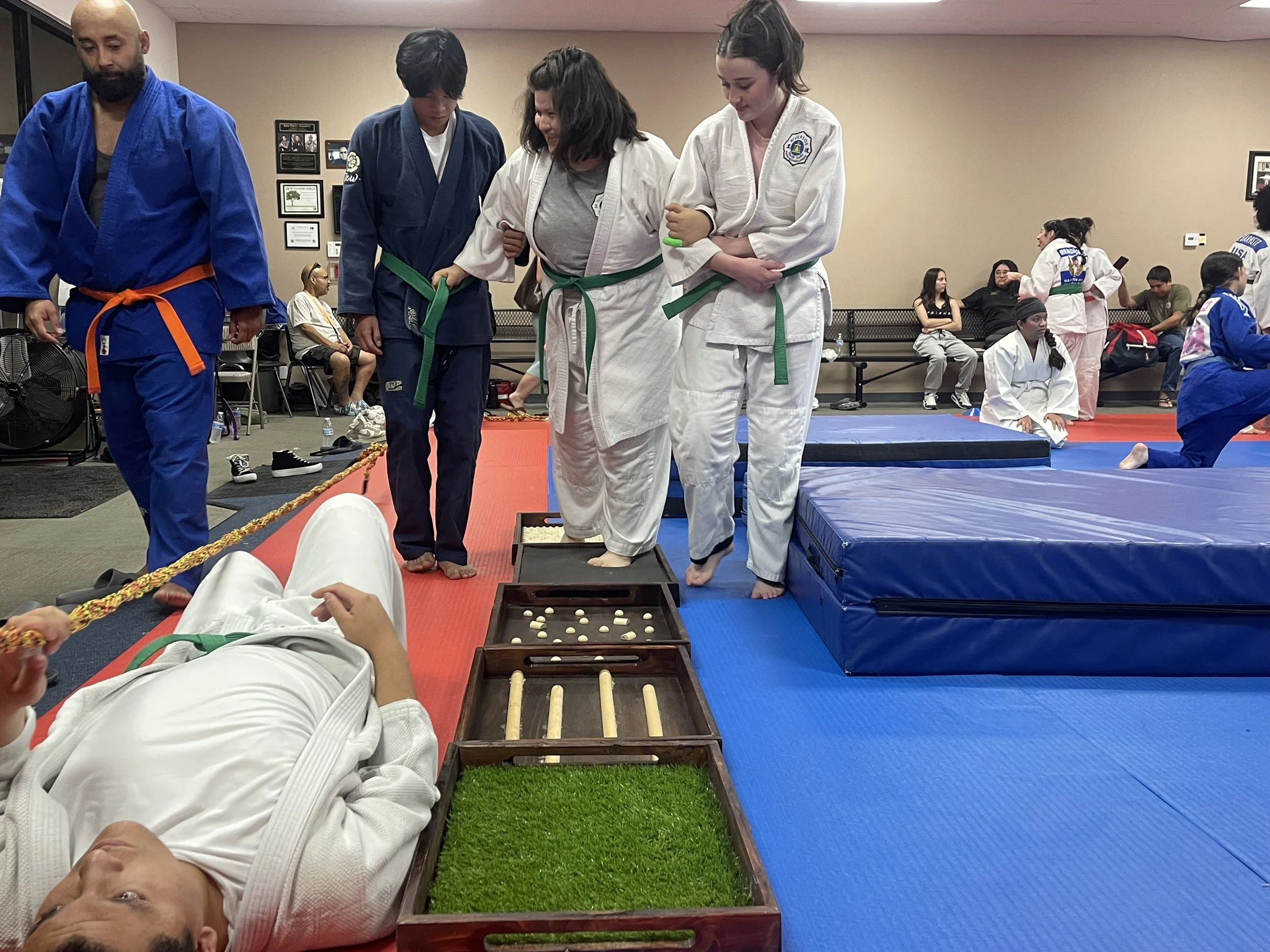 A person in a white judo gi lying on the floor with green belt, surrounded by friends in judo gi, some standing on a platform, others sitting on the mats, in a martial arts dojo setting.