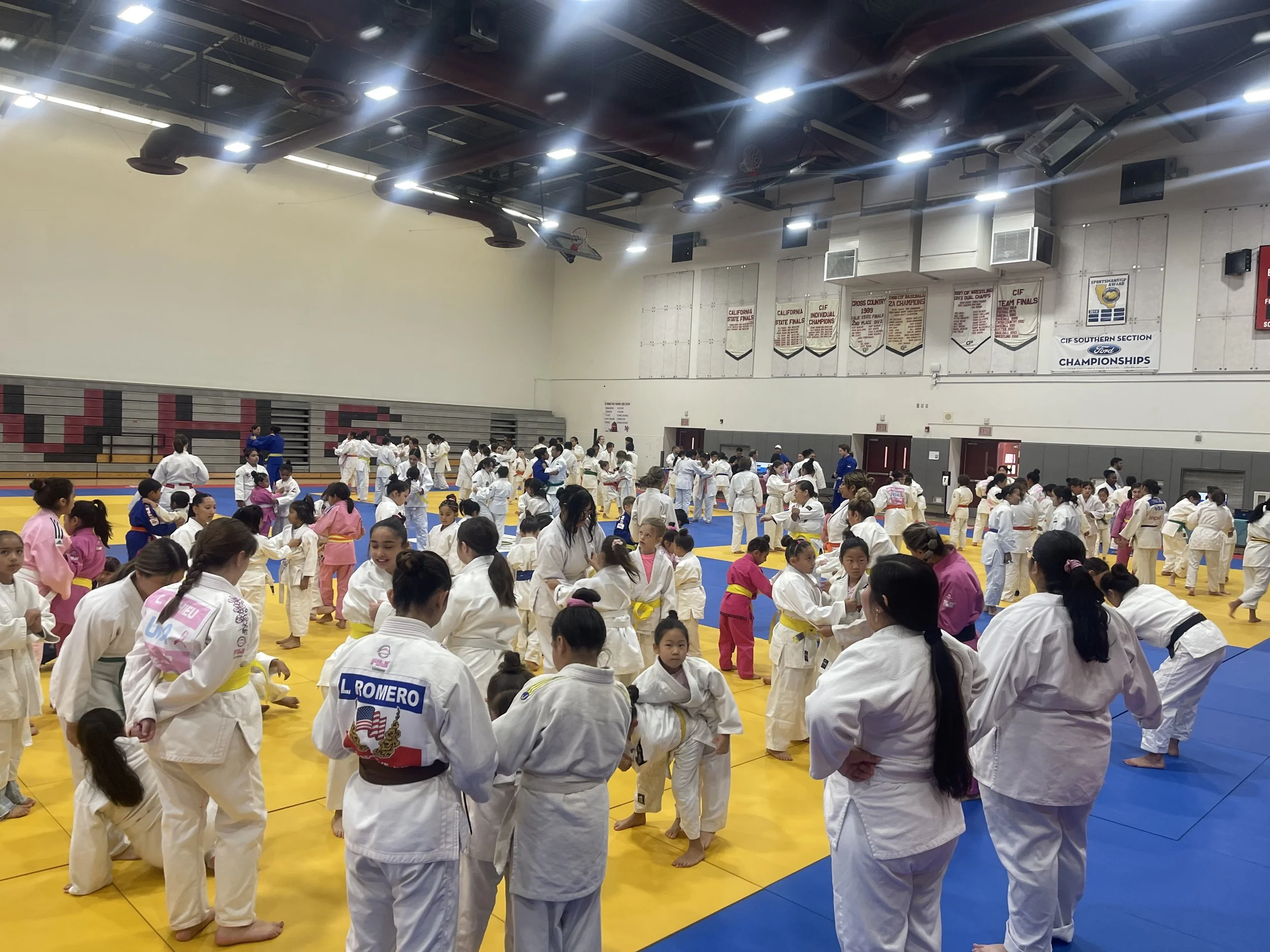 Large indoor judo competition with children and adults practicing on yellow and blue mats, some in pink gis, in a gymnasium with banners hanging on the walls.