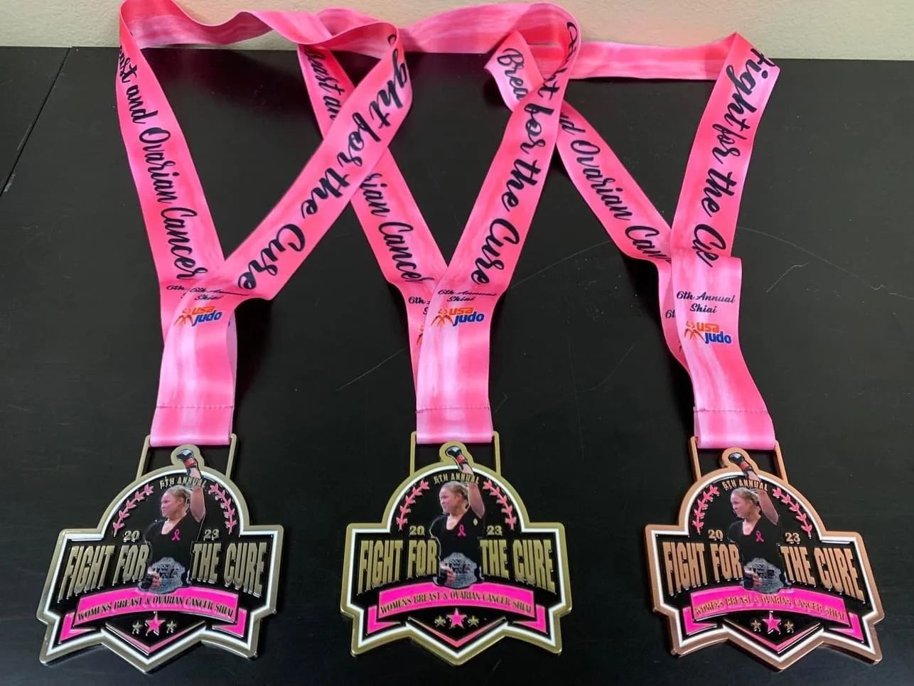 Three medals for a breast cancer awareness event, each with a pink ribbon and a photo of a woman, attached to pink ribbons with black text reading "Fight for the Cure" and "Women's Breast & Ovarian Cancer Show." The ribbons also have a logo that read
