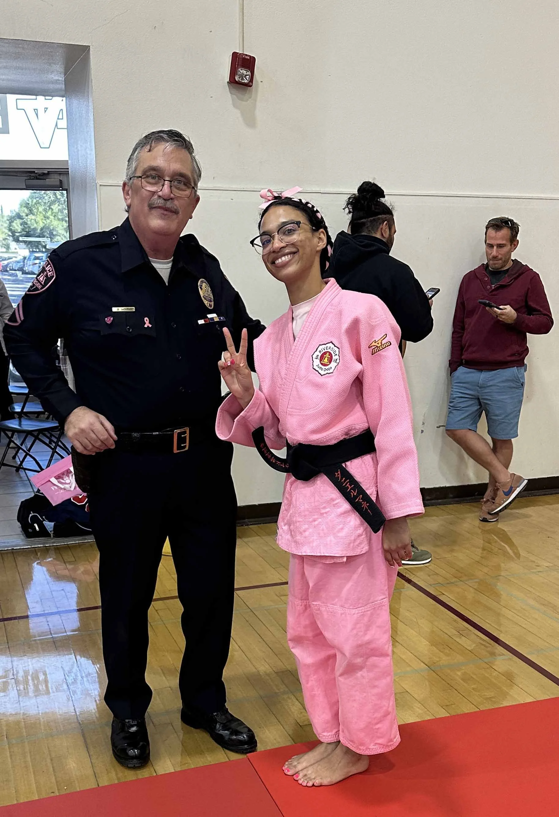 A police officer and a woman in a pink judo uniform posing together indoors, with the woman making a peace sign. Two other people are in the background, one using a phone and the other looking at a device.