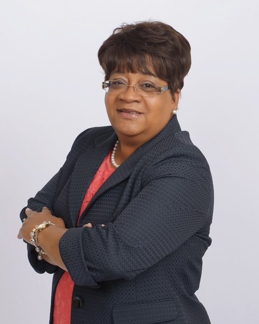 A confident middle-aged African American woman with short hair, wearing glasses, a navy blazer, pearl necklace, and bracelet, crossing her arms and smiling at the camera against a plain white background.