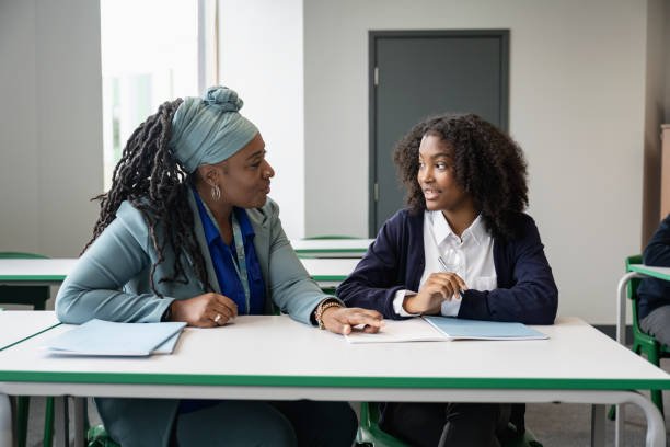 A teacher and student sitting at a desk in a classroom, engaged in conversation.