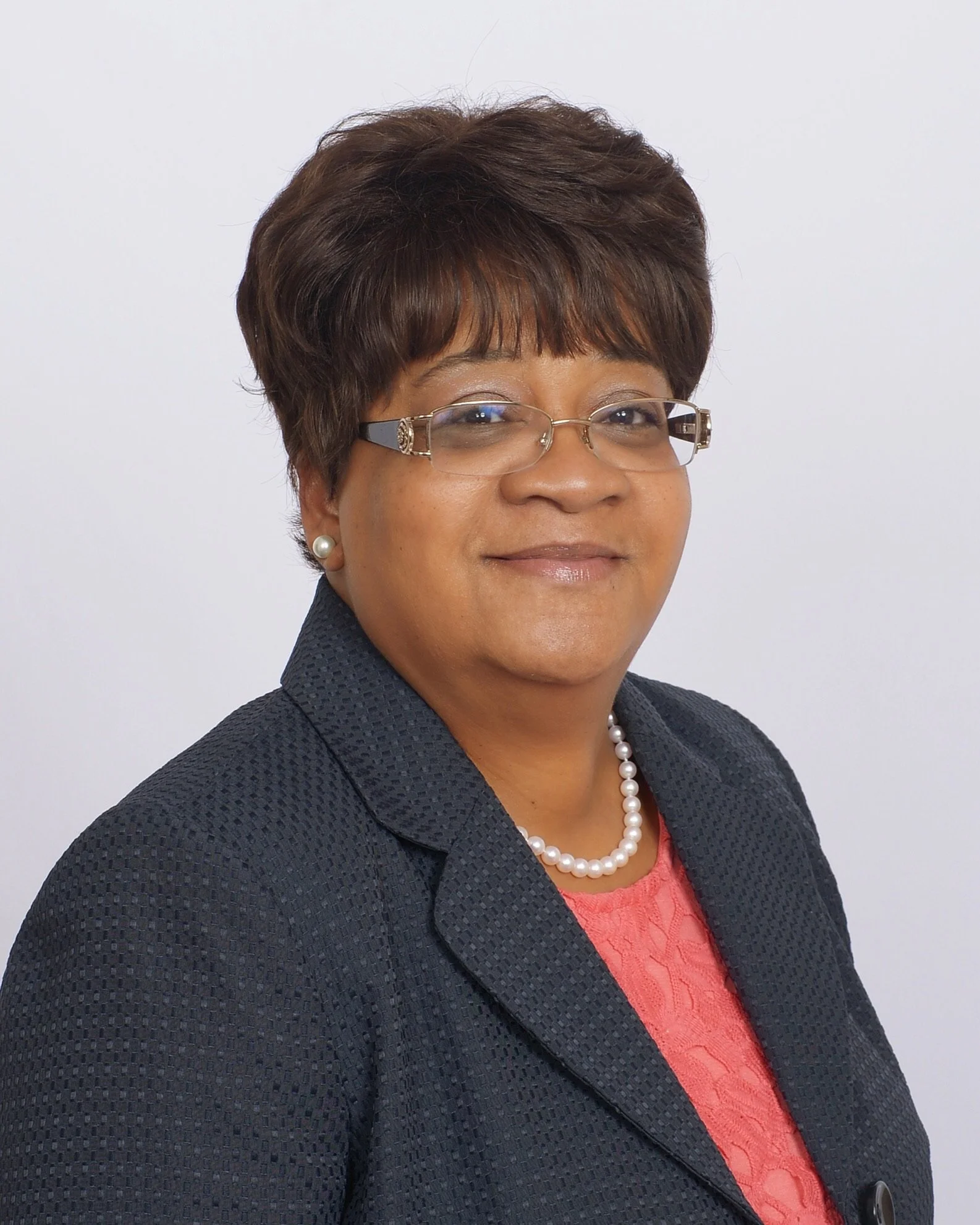 Portrait of a smiling woman with short brown hair, glasses, pearl earrings, a pearl necklace, wearing a dark blazer over a pink top, against a white background.