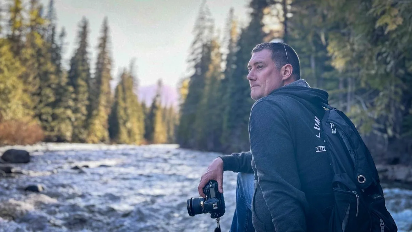A man sitting by a river in a forest, holding a camera with trees and mountain in the background.