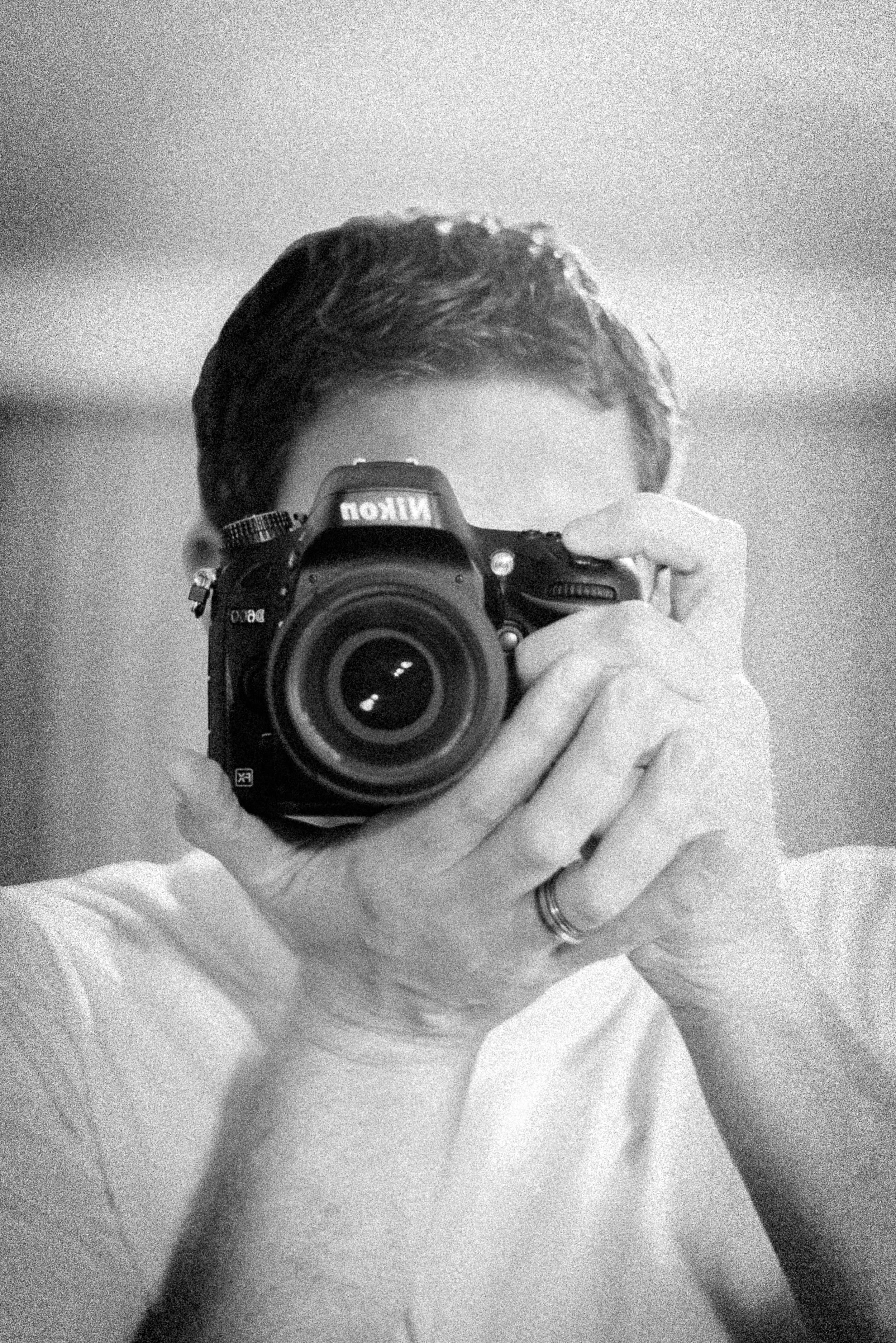 A person taking a selfie with a DSLR camera, partially obscuring their face, in a grayscale photo.