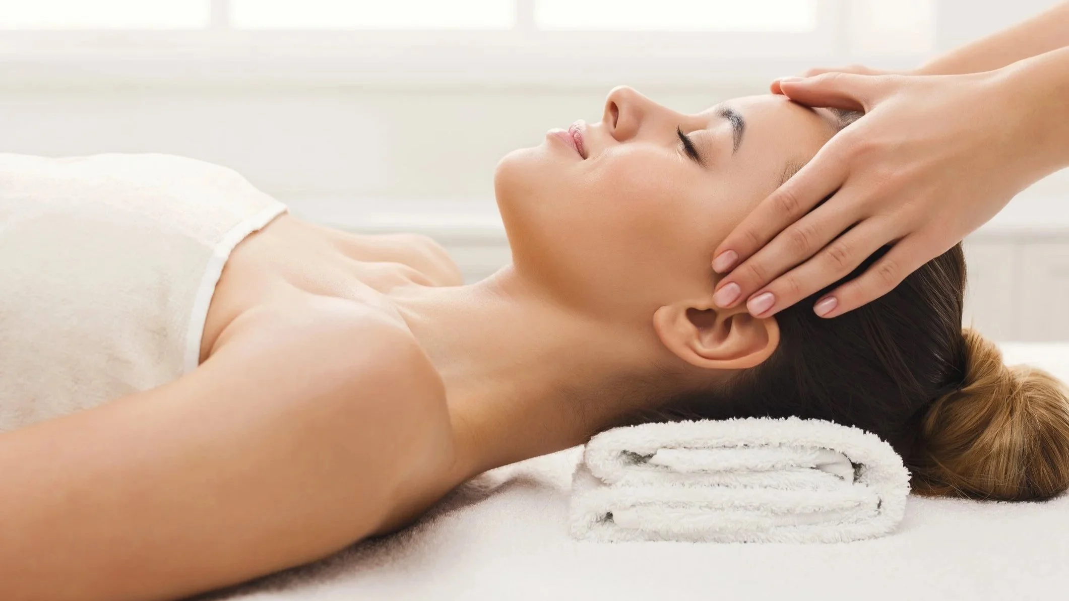 A woman receiving a head massage at a spa, lying on a white towel with her eyes closed.