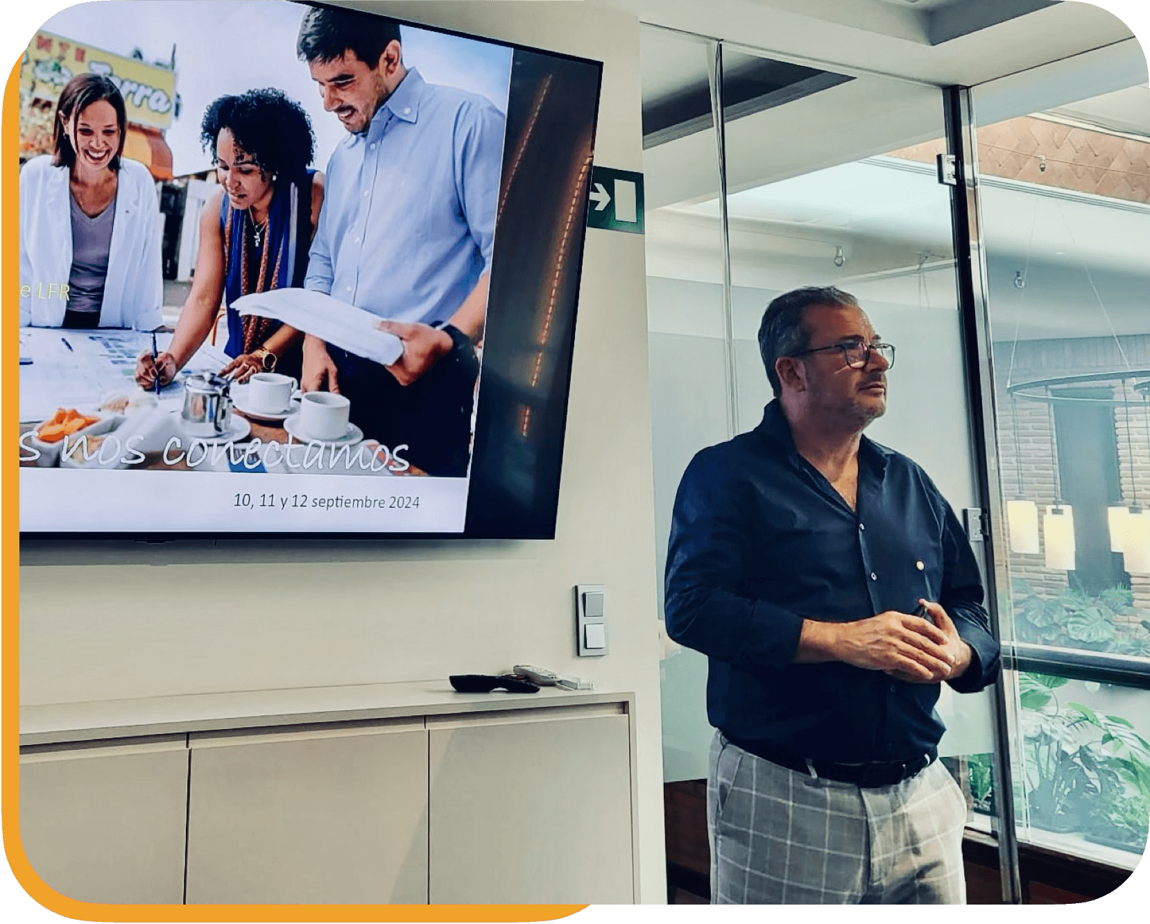 Hombre de cabello gris y barba, con gafas, vestido con camisa negra y pantalones a cuadros, observando una pantalla en una oficina moderna con ventanas de acrílico y plantas en el exterior.