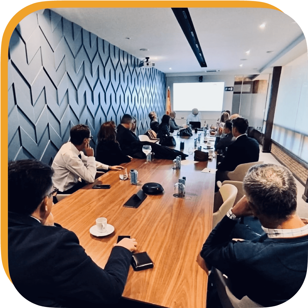 Reunión de negocios en una sala moderna con varias personas sentadas alrededor de una mesa larga, con una pantalla en la pared mostrando una presentación y bebidas en la mesa.