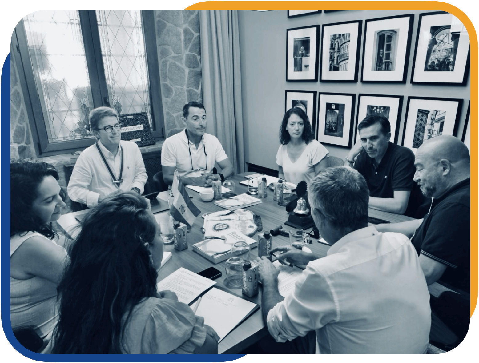 Reunión de varias personas en una sala de conferencias, sentadas alrededor de una mesa con documentos y bebidas, en un entorno con paredes de piedra y fotografías enmarcadas.
