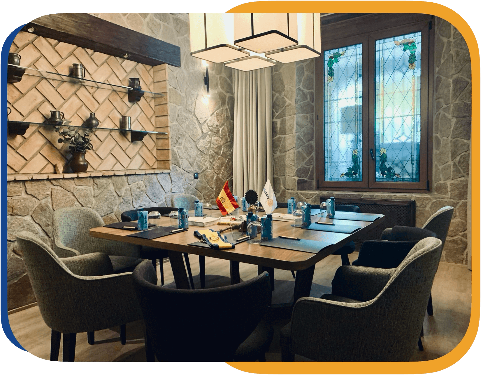 Sala de reuniones con mesa rectangular y sillas acolchadas, paredes de piedra, ventana con vitrales decorativos y estantería de ladrillo con decoraciones metálicas.