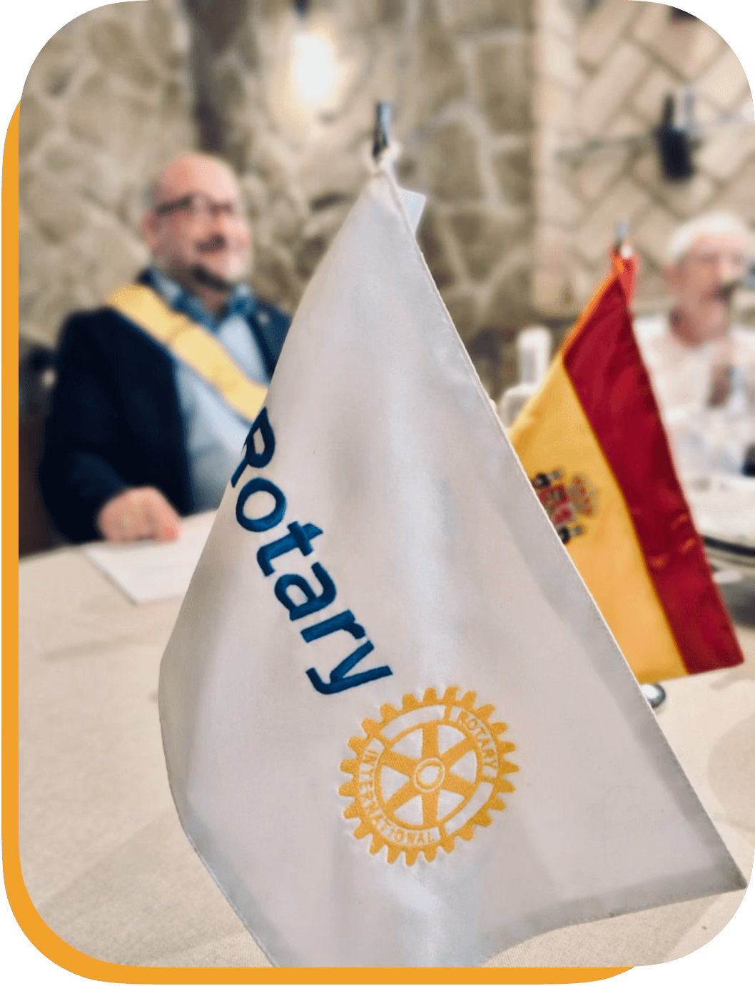 Banderas en una mesa, una con el texto 'Rotary' y el símbolo del Rotary International, y otra bandera de España, con fondo borroso de personas en una reunión.