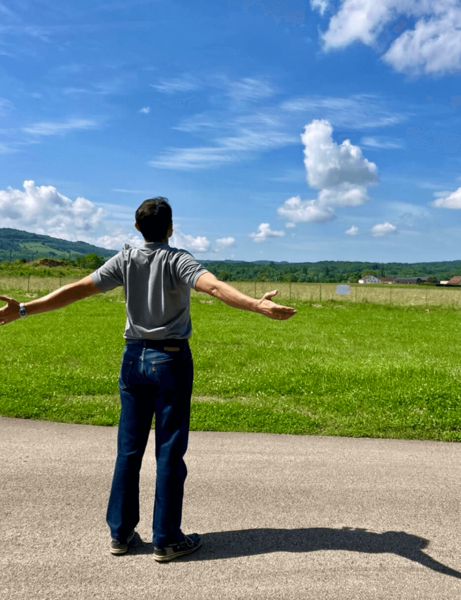 Hombre de espaldas con los brazos extendidos en un campo verde, cielo azul con nubes blancas y algunos edificios y colinas en la distancia.