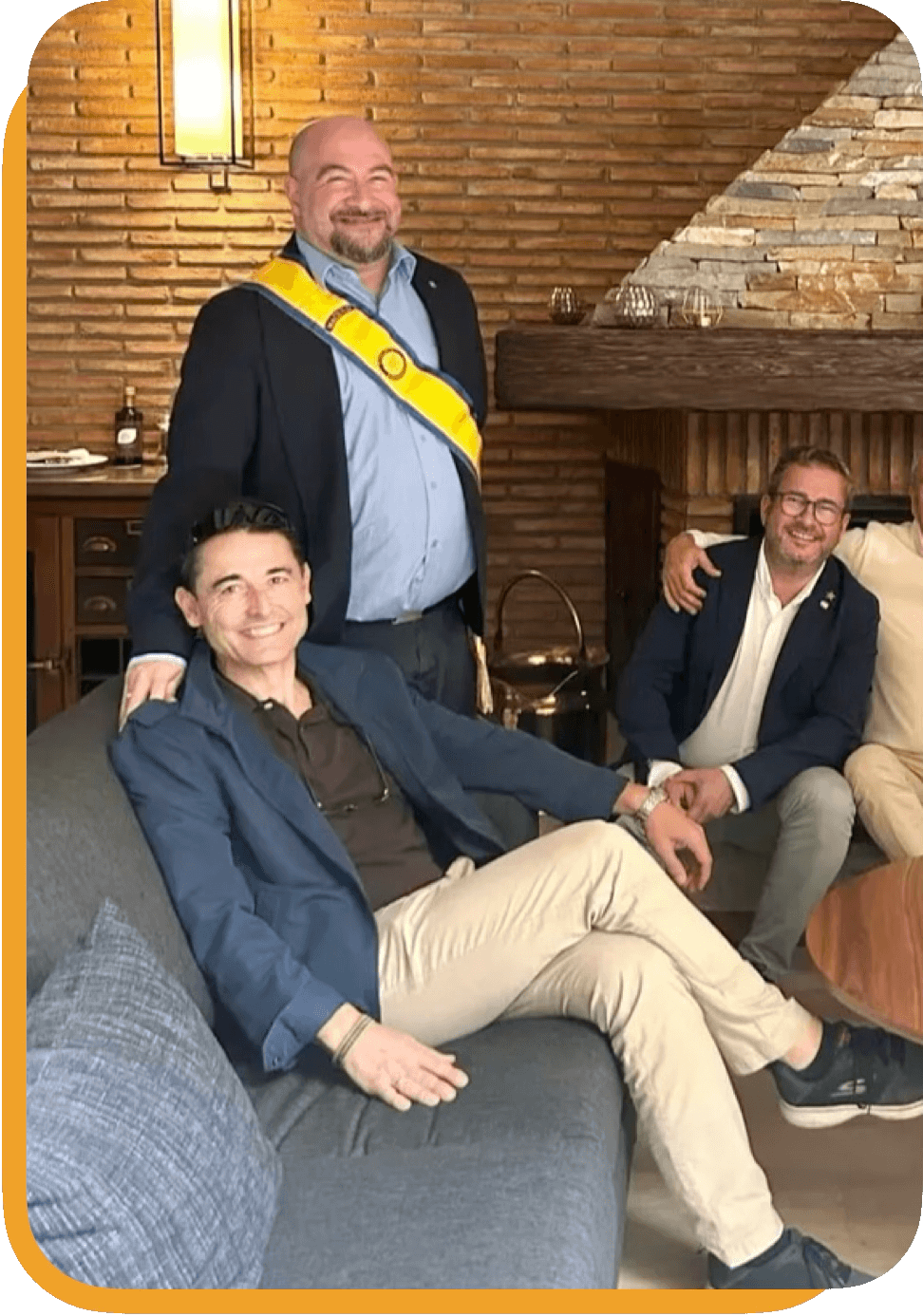 Grupo de cuatro hombres en una reunión social en una sala con paredes de ladrillo y una chimenea de ladrillos. Uno de los hombres en el centro usa una banda amarilla en el hombro y una chaqueta negra, y está de pie. Los otros tres hombres están sentados en un sofá, vestidos con chaquetas y pantalones claros y oscuros, sonriendo y mostrando camaradería.