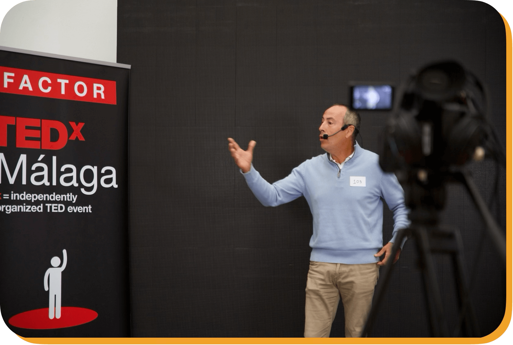 Hombre dando una charla en un evento TEDx en Málaga, usando un micrófono de diadema y un gafete con el número 103, frente a un cartel que indica el evento TEDx en Málaga.