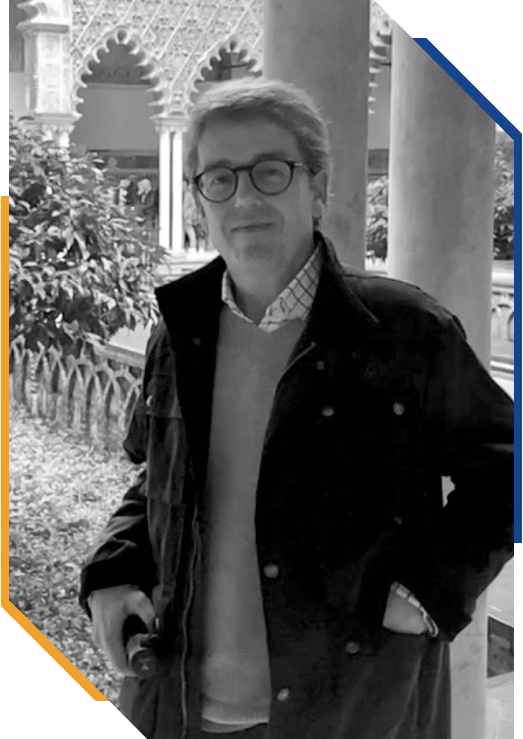 Hombre con gafas de sol, chaqueta y suéter, de cabello gris y rubio, de pie frente a columnas y arbustos en un lugar con arquitectura clásica, en color negro y blanco.