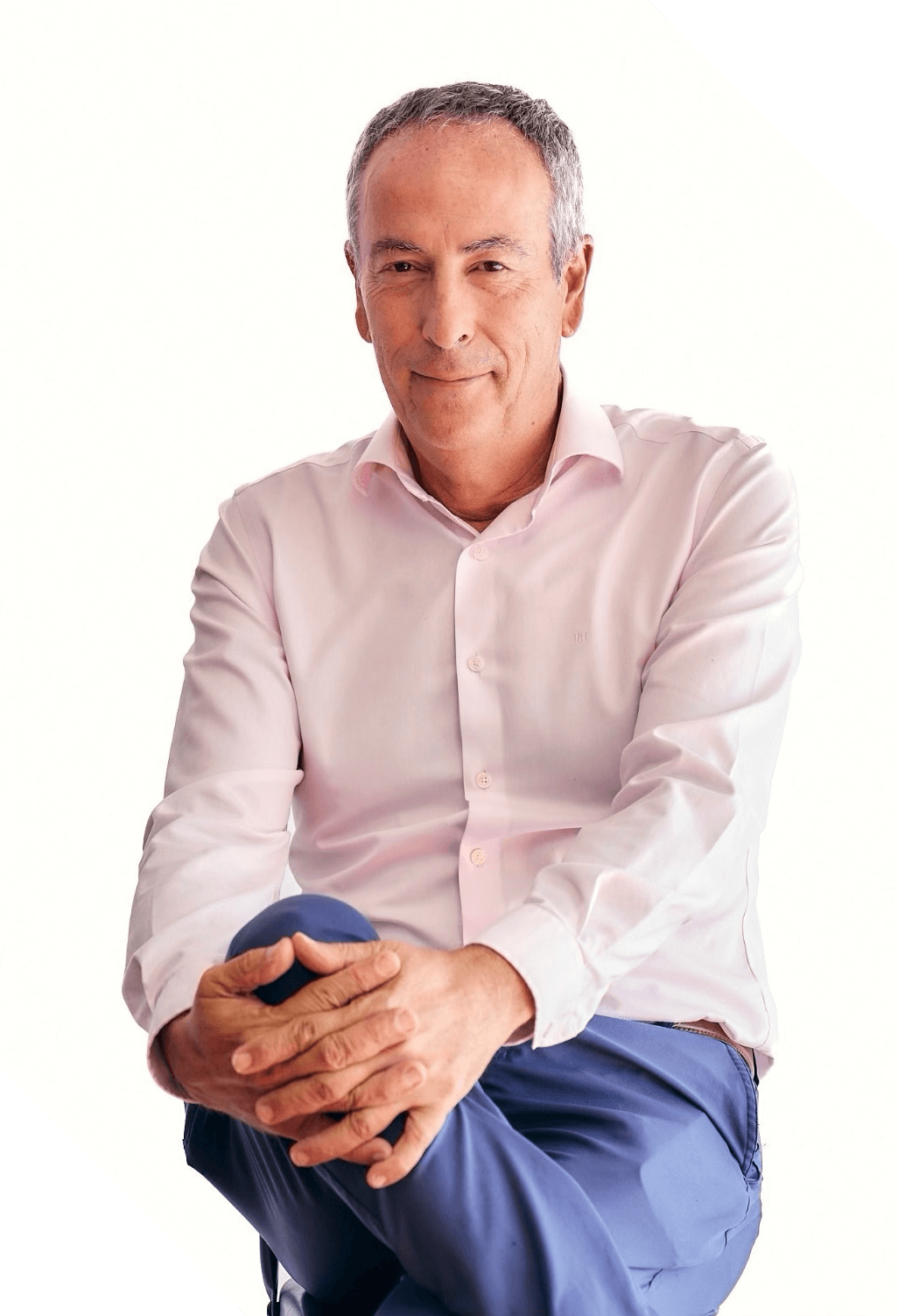 Hombre con camisa rosada y pantalones azul oscuro, sentado con las manos juntas, en un fondo blanco con esquinas negras.