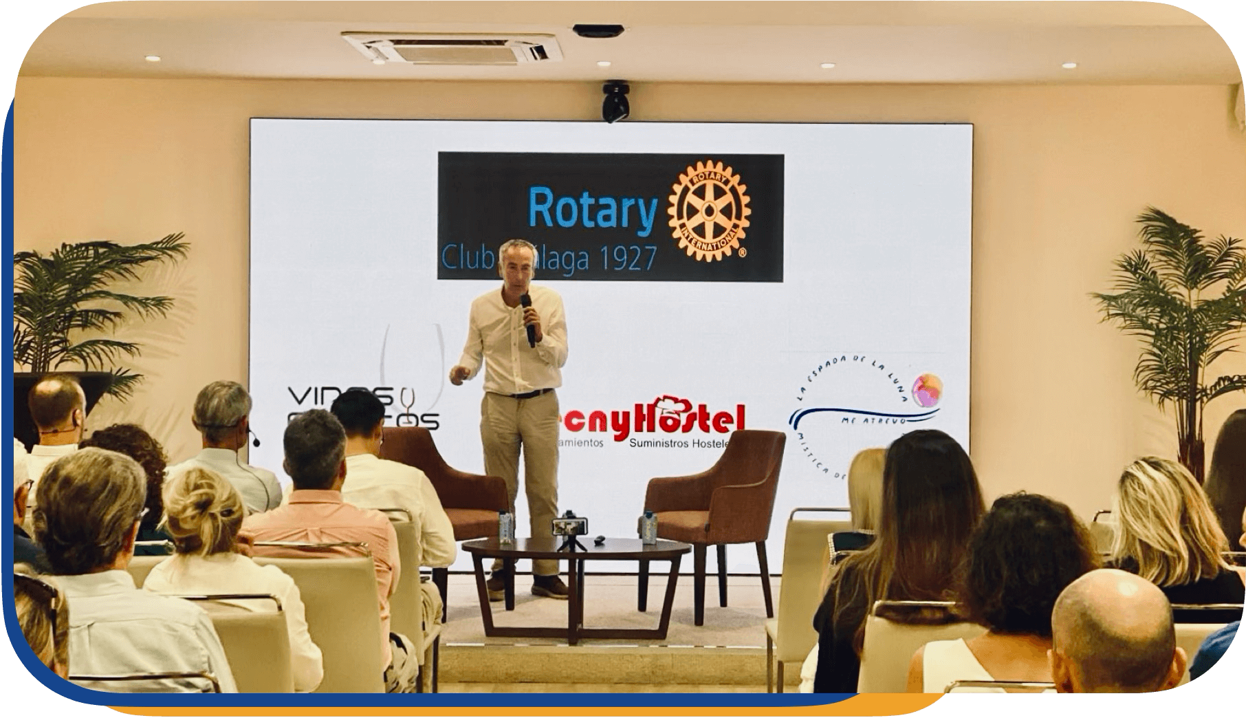 Hombre en escenario presentando frente a audiencia en evento del Rotary Club en Málaga, España, con pantalla que muestra logos y texto en español.