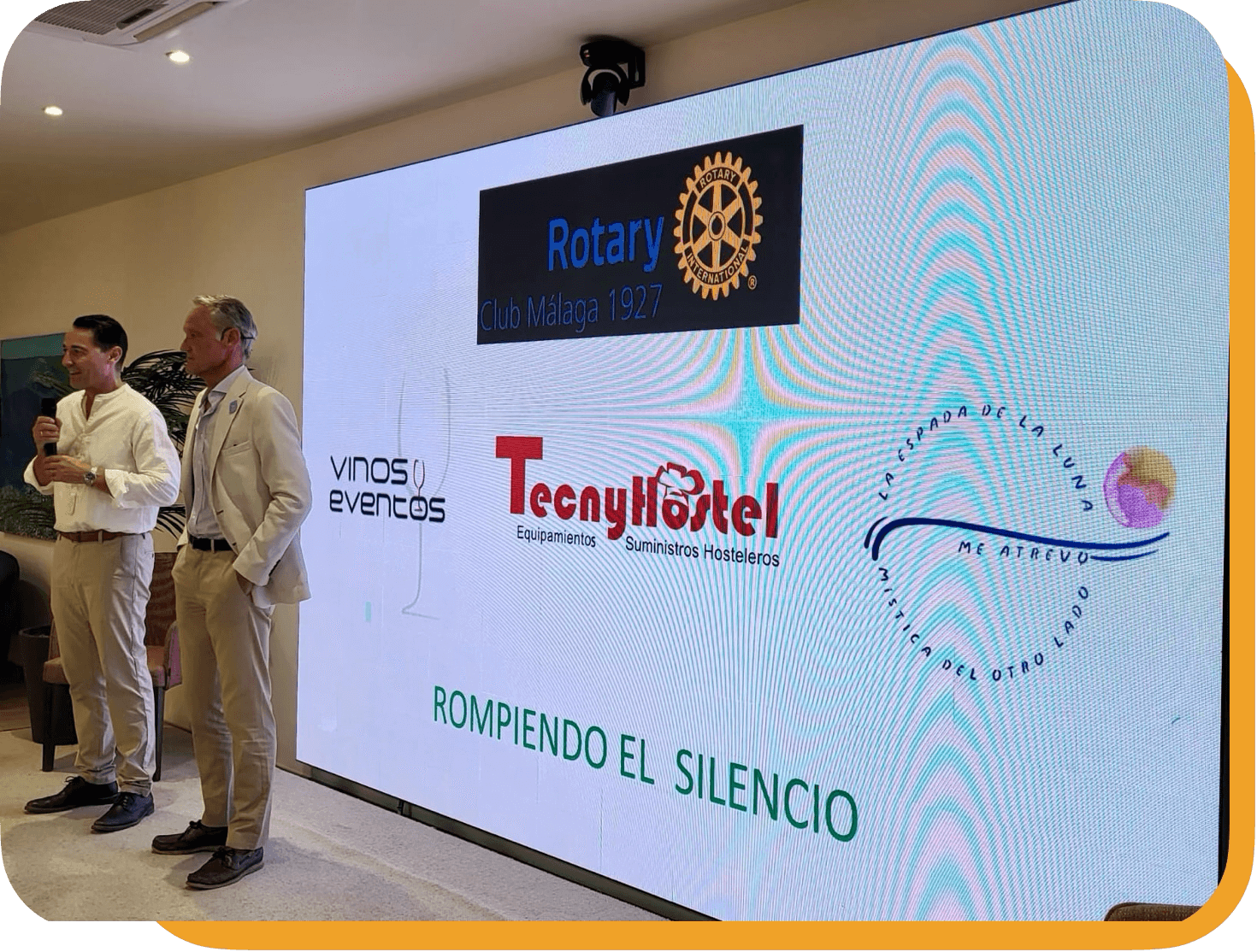 Una pantalla grande con varios logotipos y textos, al fondo hay dos hombres en el escenario en una presentación o evento. En la pantalla se ven logos de Rotary Club Málaga, Vinos y Eventos, TecnayHostel y un diseño con el texto 'La Esparada de La Luna, Me Atrevo, Con Otro Lado'. Abajo, en letras verdes, dice 'ROMPIENDO EL SILENCIO'.