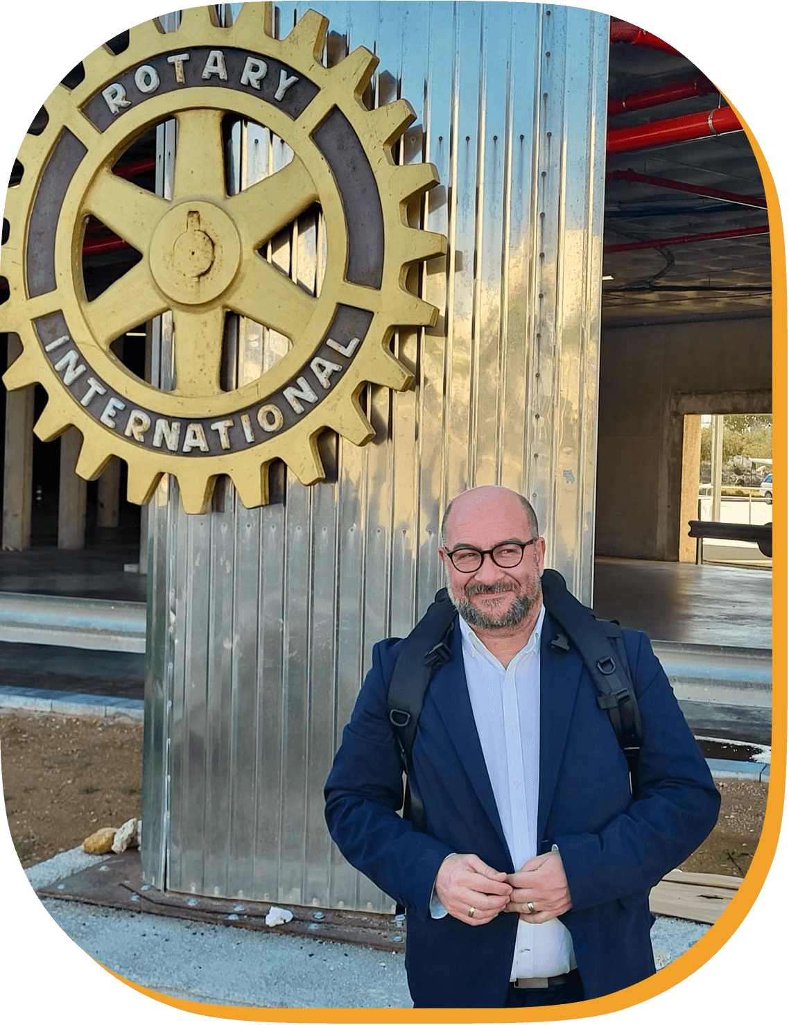 Hombre con gafas y barba, usando chaqueta azul y mochila, sonriendo frente a un cartel grande de engranaje con la inscripción 'Rotary International' en un edificio en construcción.