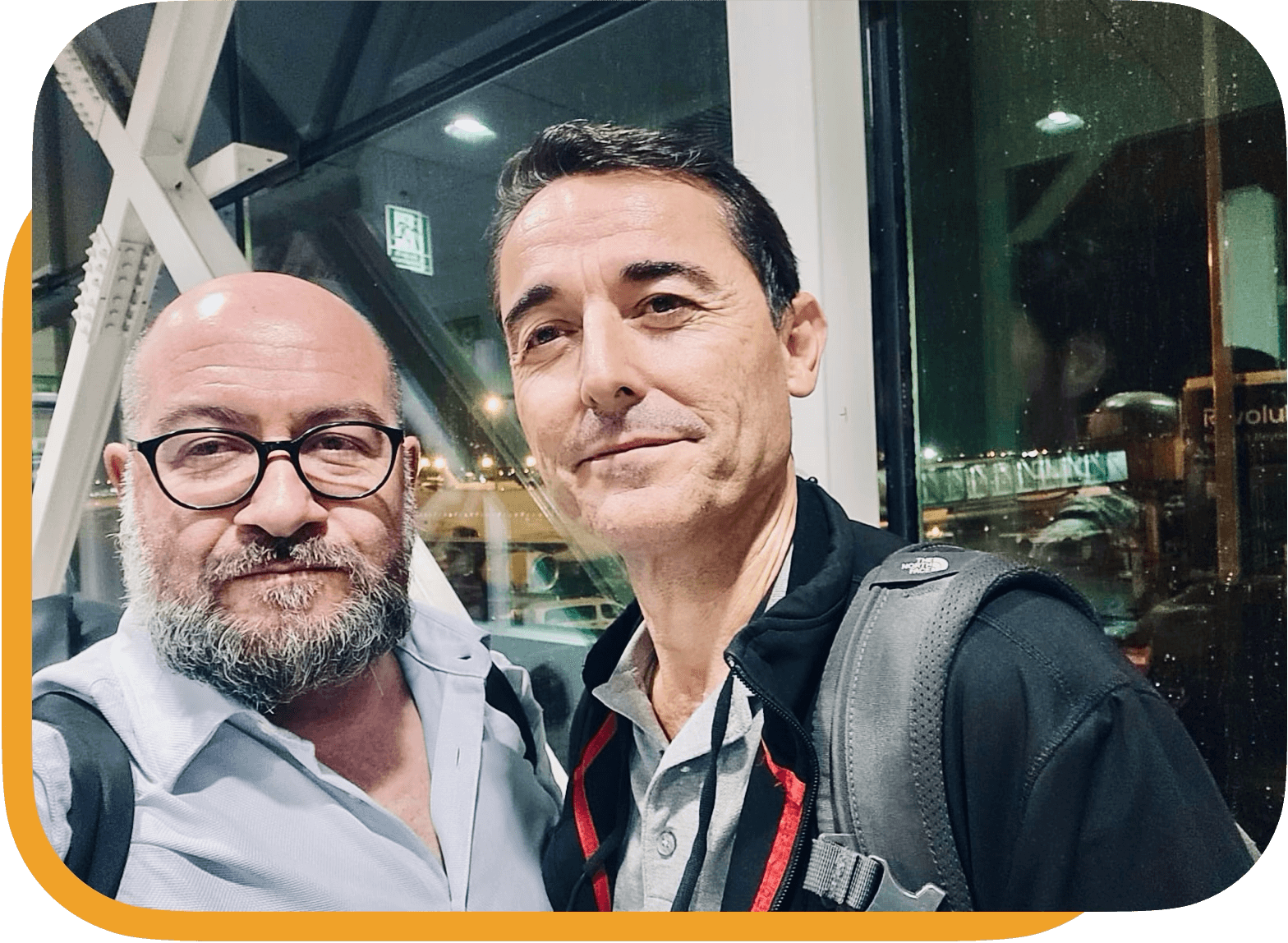Dos hombres en un aeropuerto, uno con gafas y barba, y otro con cabello oscuro y mochila, posando para una selfie.