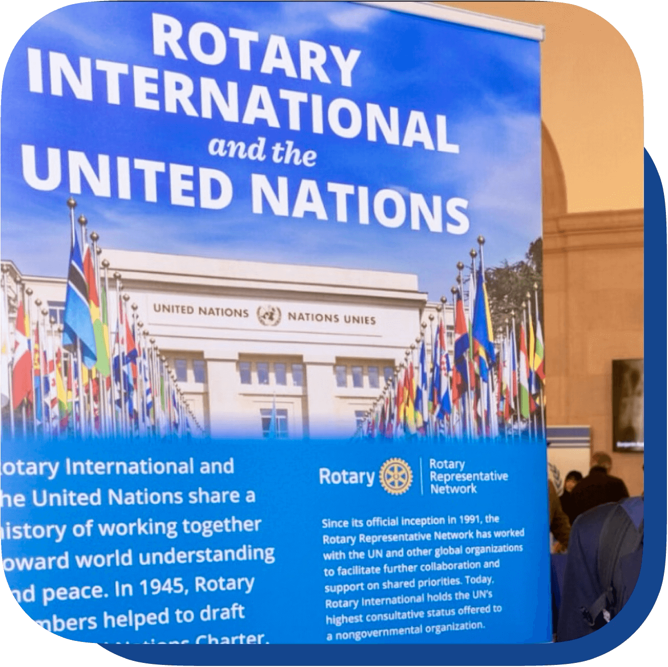 Pancarta sobre Rotary Internacional y las Naciones Unidas, en un evento con varias banderas internacionales, en un interior con gente observando.